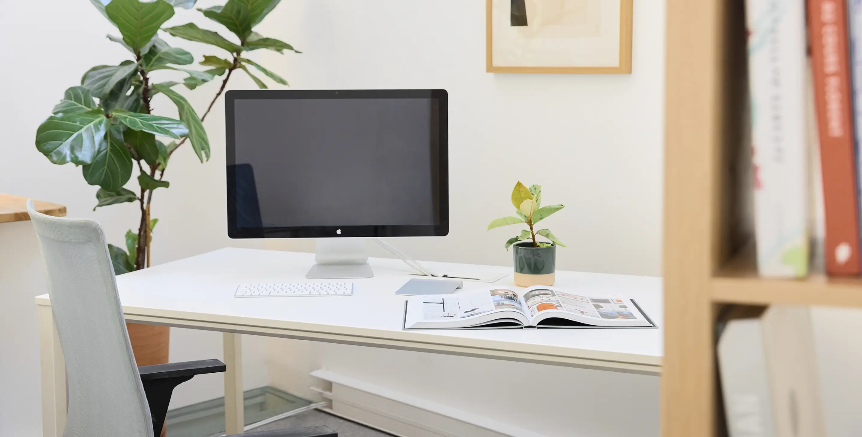 Bureau moderne avec écran d'ordinateur éteint, clavier, plante en pot et magazine ouvert sur une table blanche, avec une grande plante verte en arrière-plan.