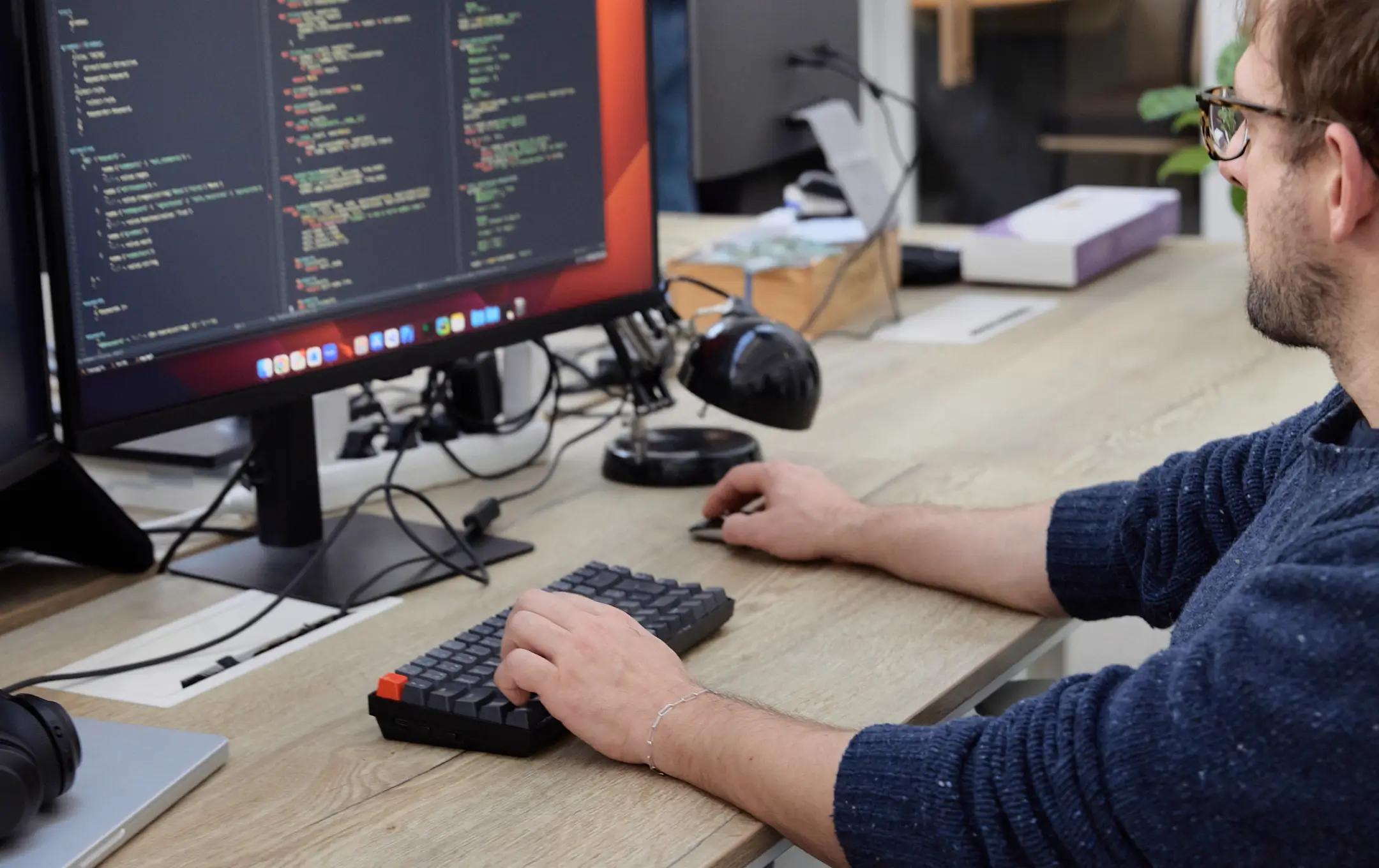 Homme portant un pull bleu travaillant sur un ordinateur avec un écran affichant du code, utilisant un clavier compact et une souris sur un bureau en bois.