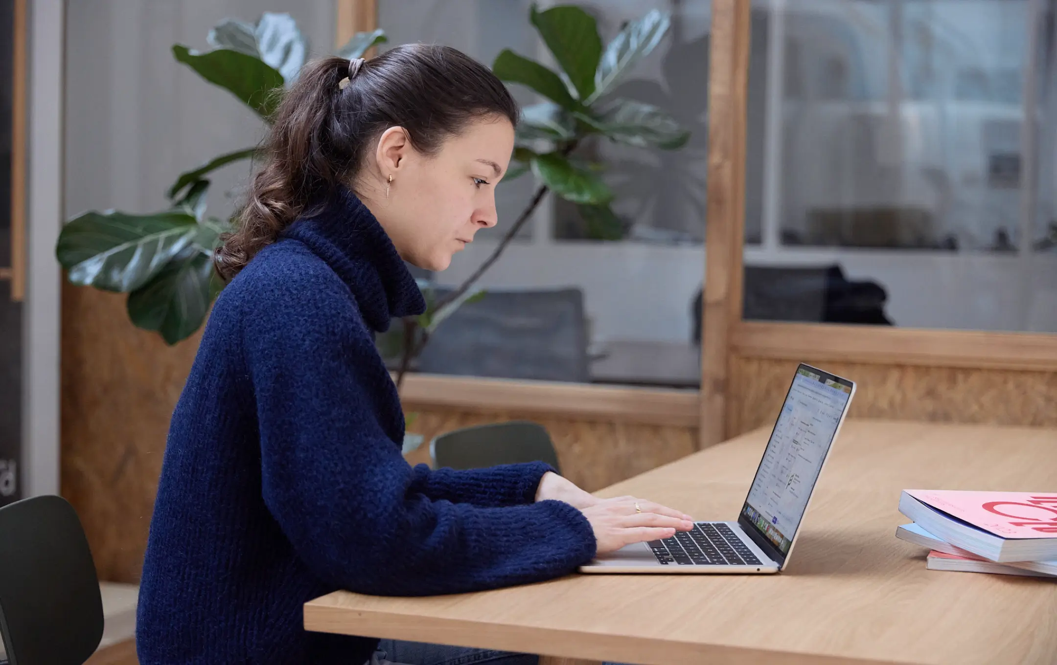 Jeune femme en pull bleu travaillant sur un ordinateur portable dans un bureau moderne avec plante et livres sur une table en bois.