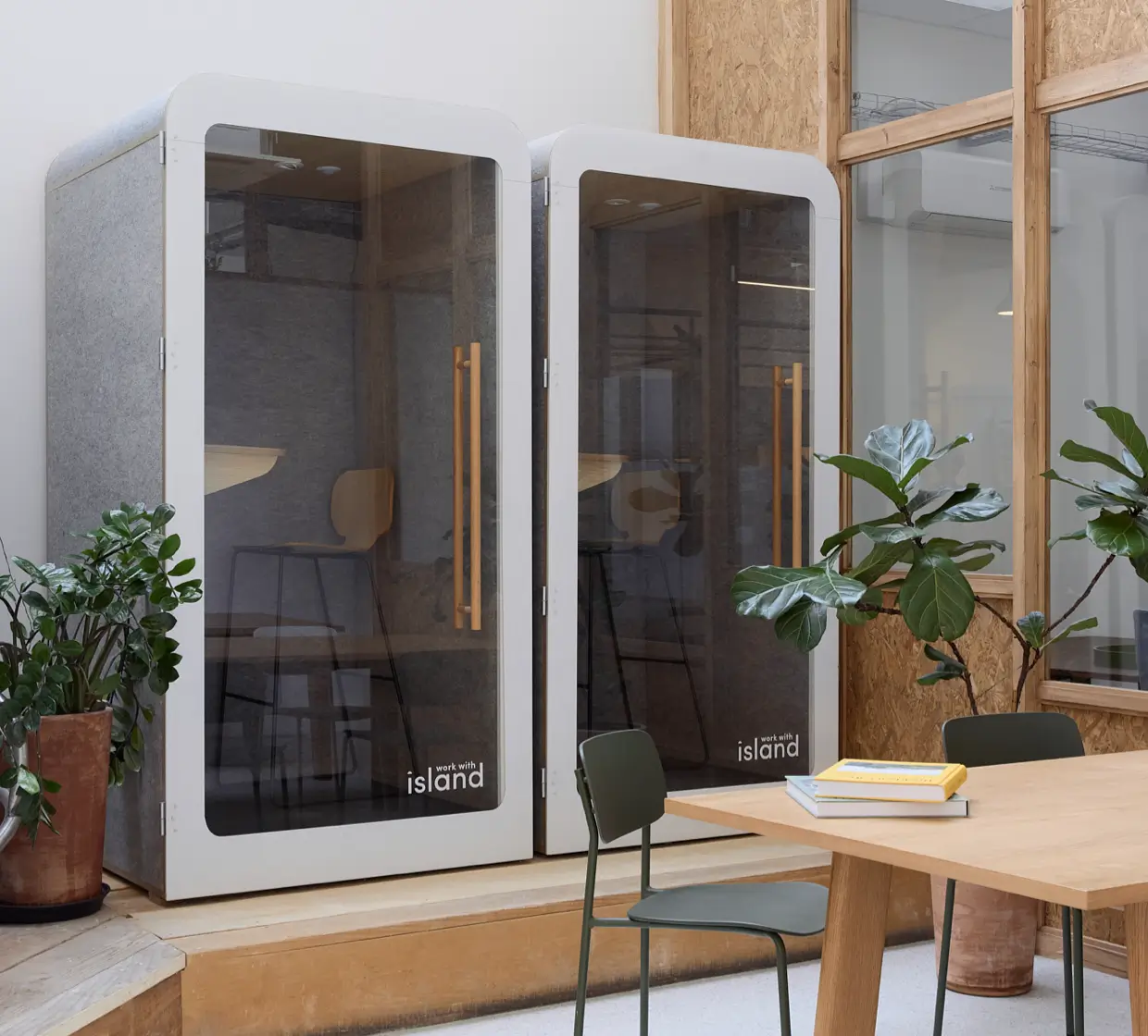 Deux cabines téléphoniques modernes en verre avec chaises hautes à l'intérieur dans un espace de travail avec plantes et table en bois.
