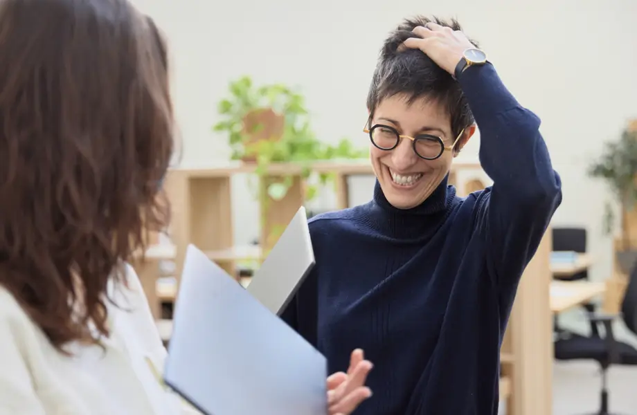 Deux femmes en conversation dans un bureau, l'une sourit en se grattant la tête tandis que l'autre tient un dossier.