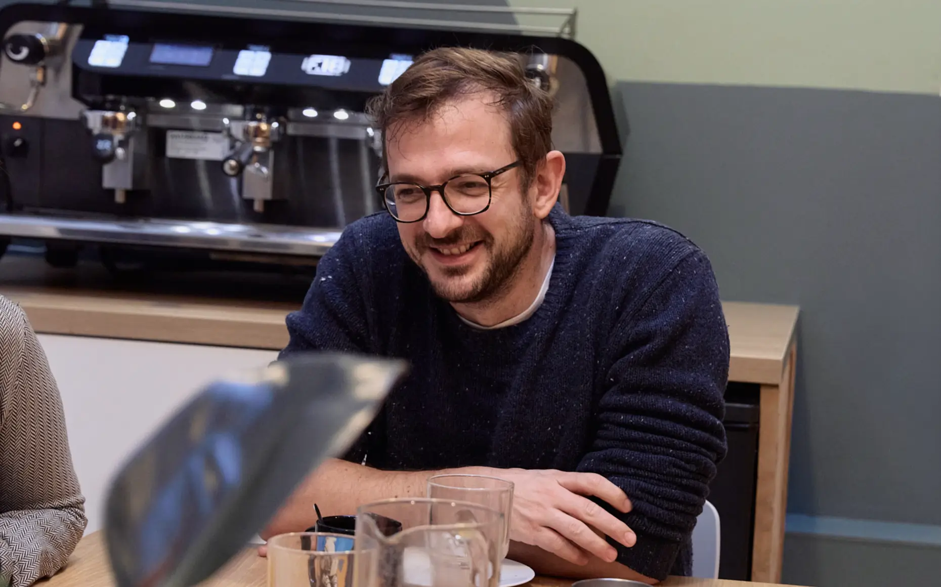 Un homme souriant en pull bleu marine assis à une table avec une machine à café en arrière-plan.
