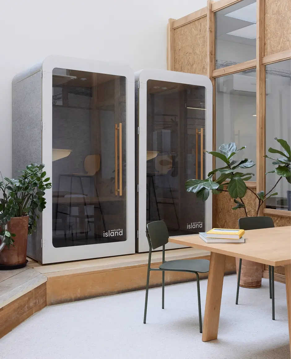 Deux cabines téléphoniques modernes avec portes vitrées et poignées en bois dans un espace de bureau avec table en bois, chaises vertes et plantes en pots.