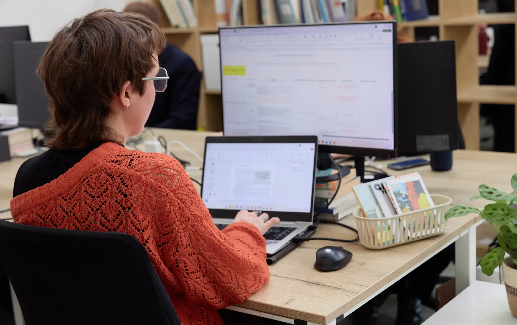 Personne au pull orange travaillant sur un ordinateur portable avec un grand écran et des livres sur un bureau en bois clair.