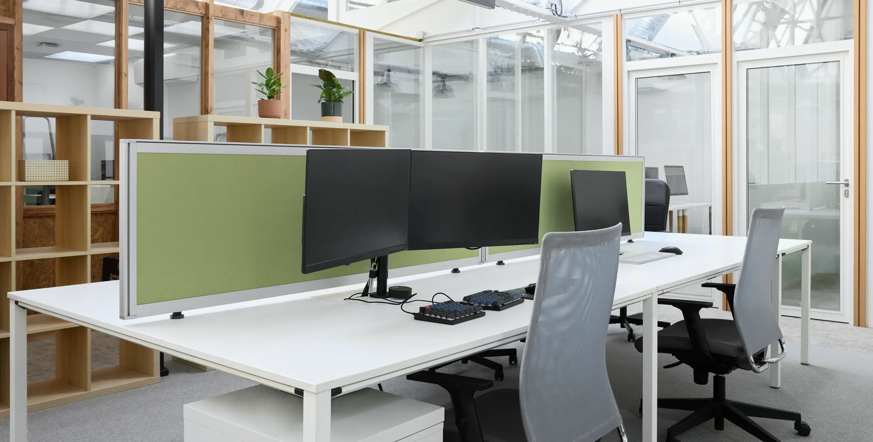 Bureau moderne avec deux stations de travail équipées d'écrans d'ordinateur, claviers mécaniques, chaises ergonomiques et séparateurs verts.