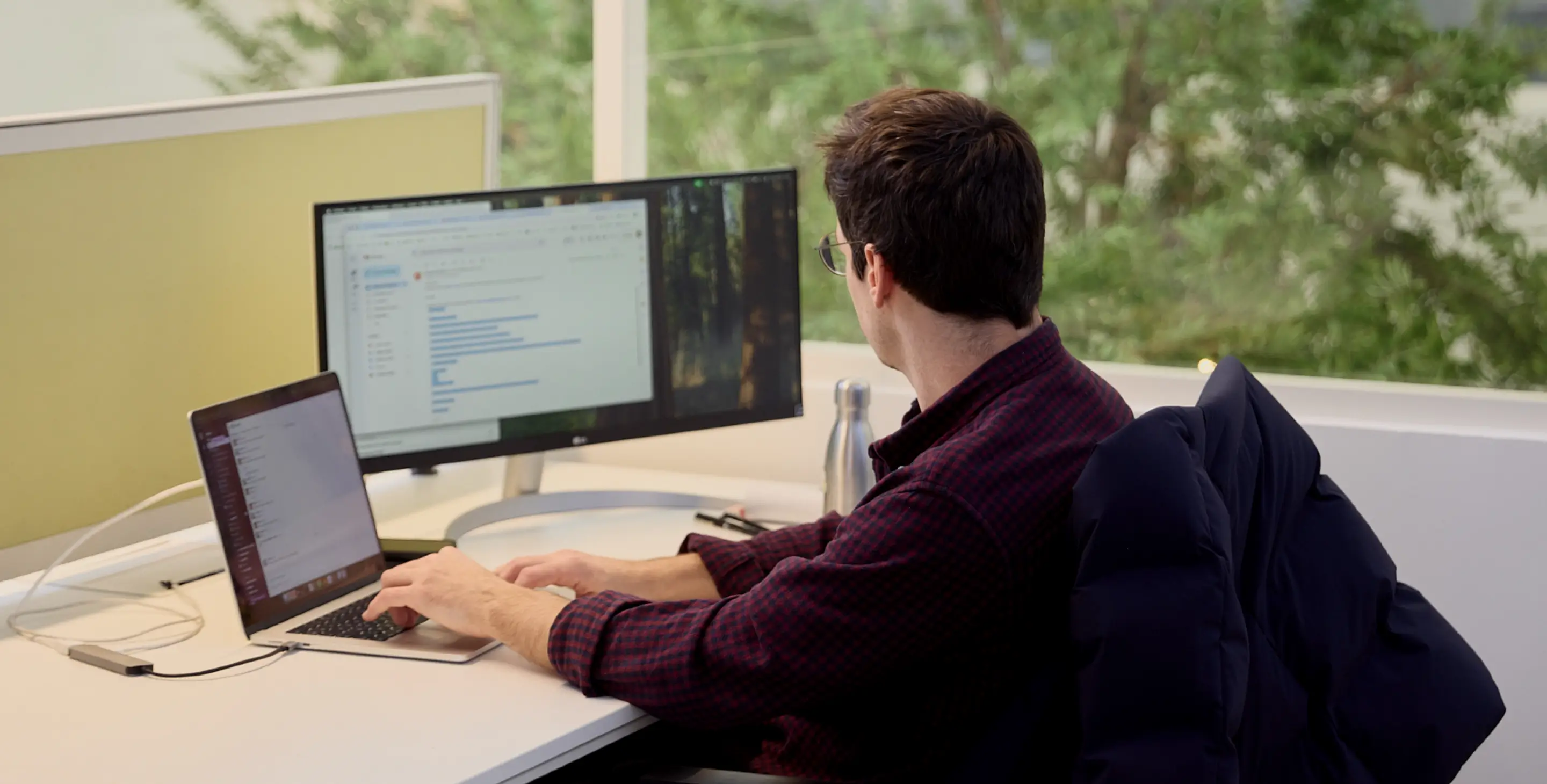 Un homme vu de dos travaille sur un ordinateur portable avec un écran supplémentaire sur un bureau près d'une fenêtre.