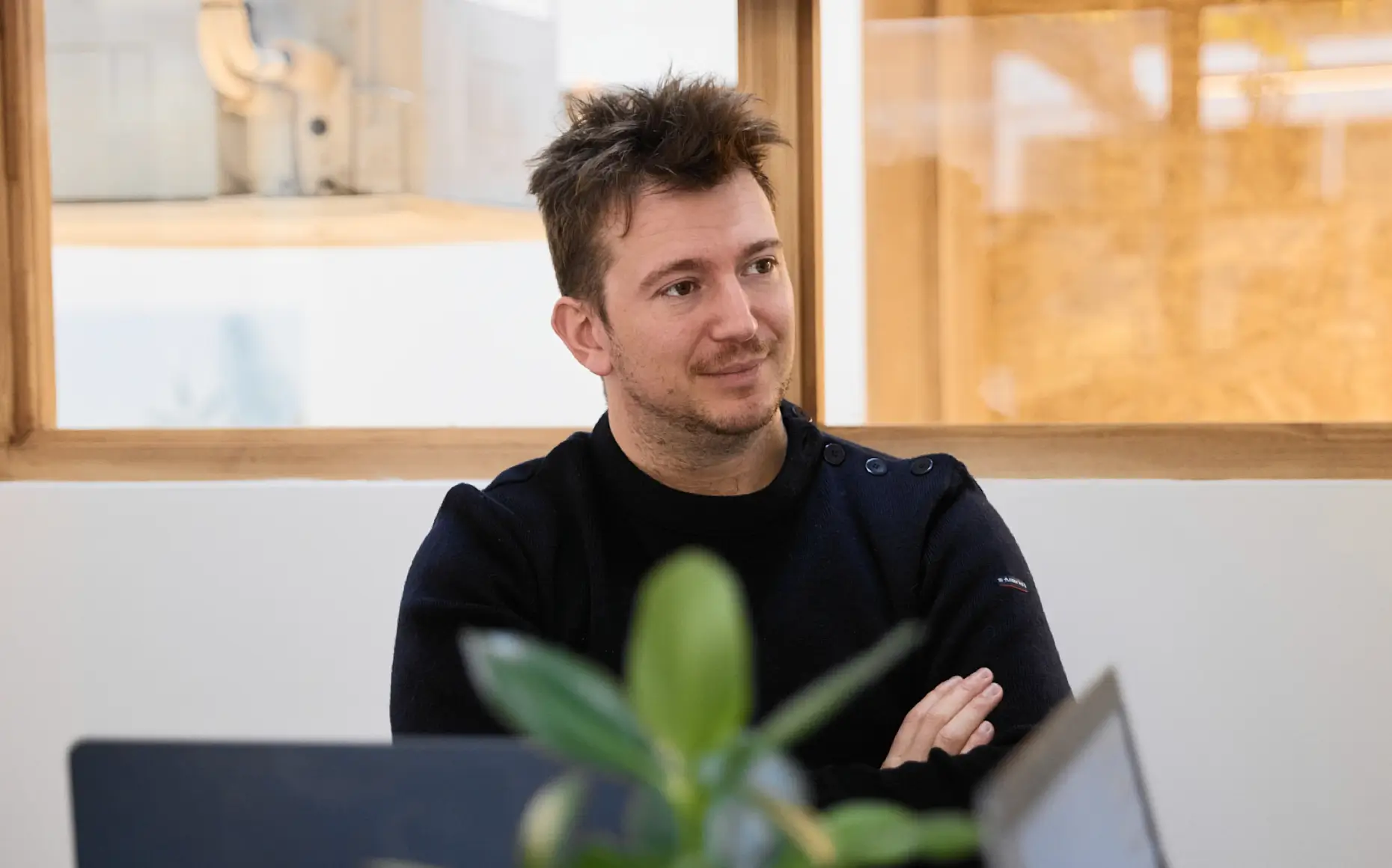 Homme avec un pull noir assis les bras croisés devant un bureau avec un ordinateur, regardant sur le côté dans une pièce lumineuse.