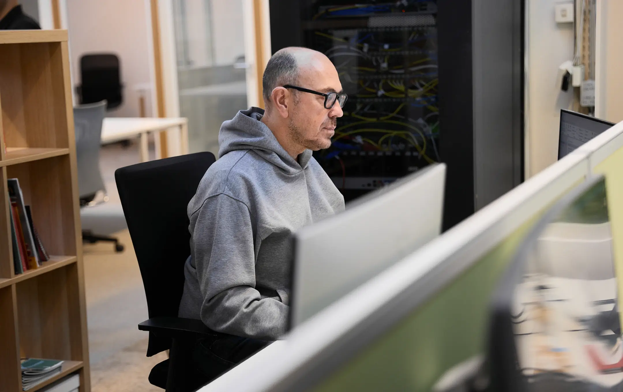 Homme portant un sweat à capuche gris et des lunettes, travaillant sur un ordinateur dans un bureau moderne avec des équipements réseau en arrière-plan.