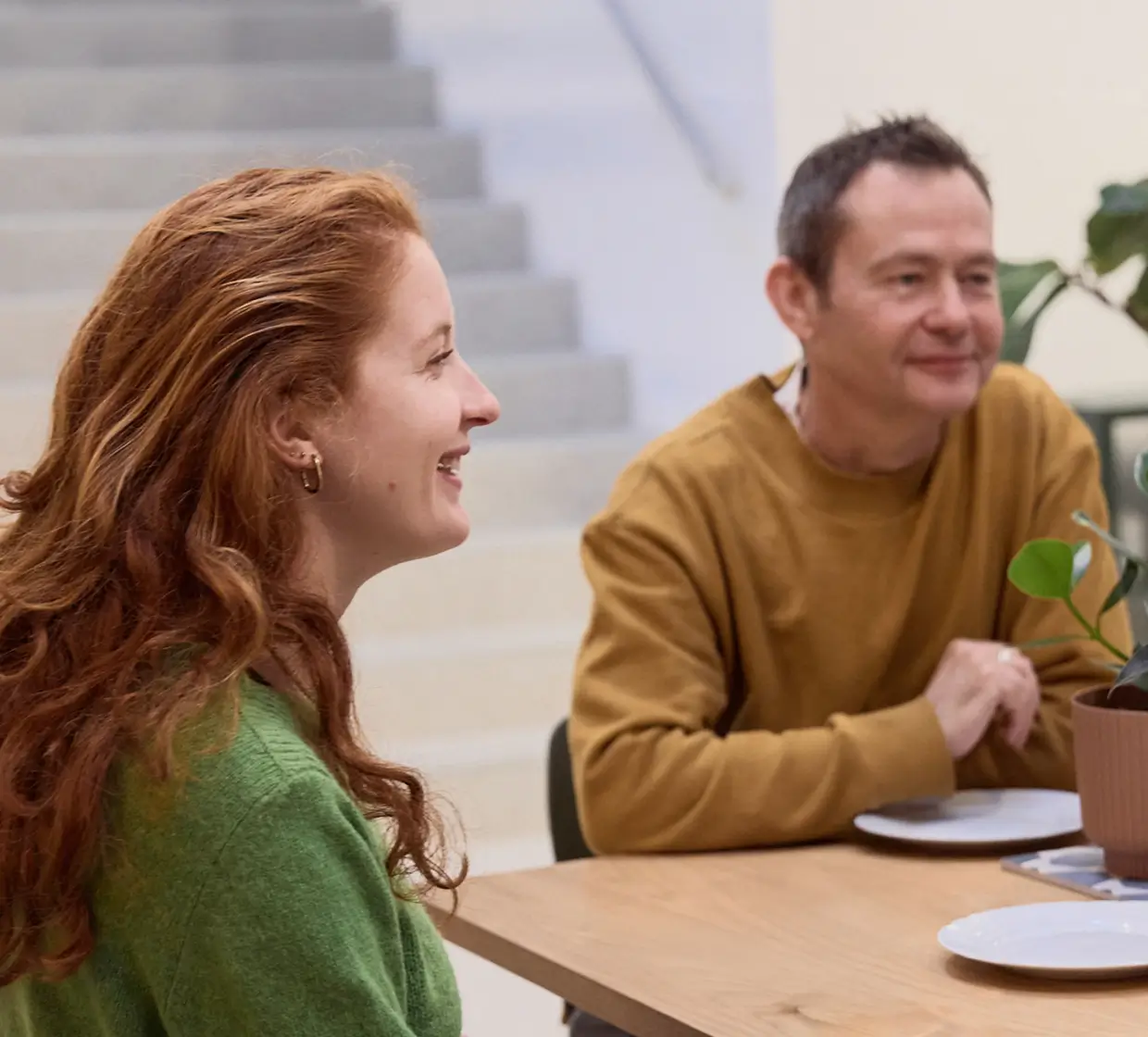 Deux personnes souriantes discutant à une table avec des assiettes vides et une plante en pot.