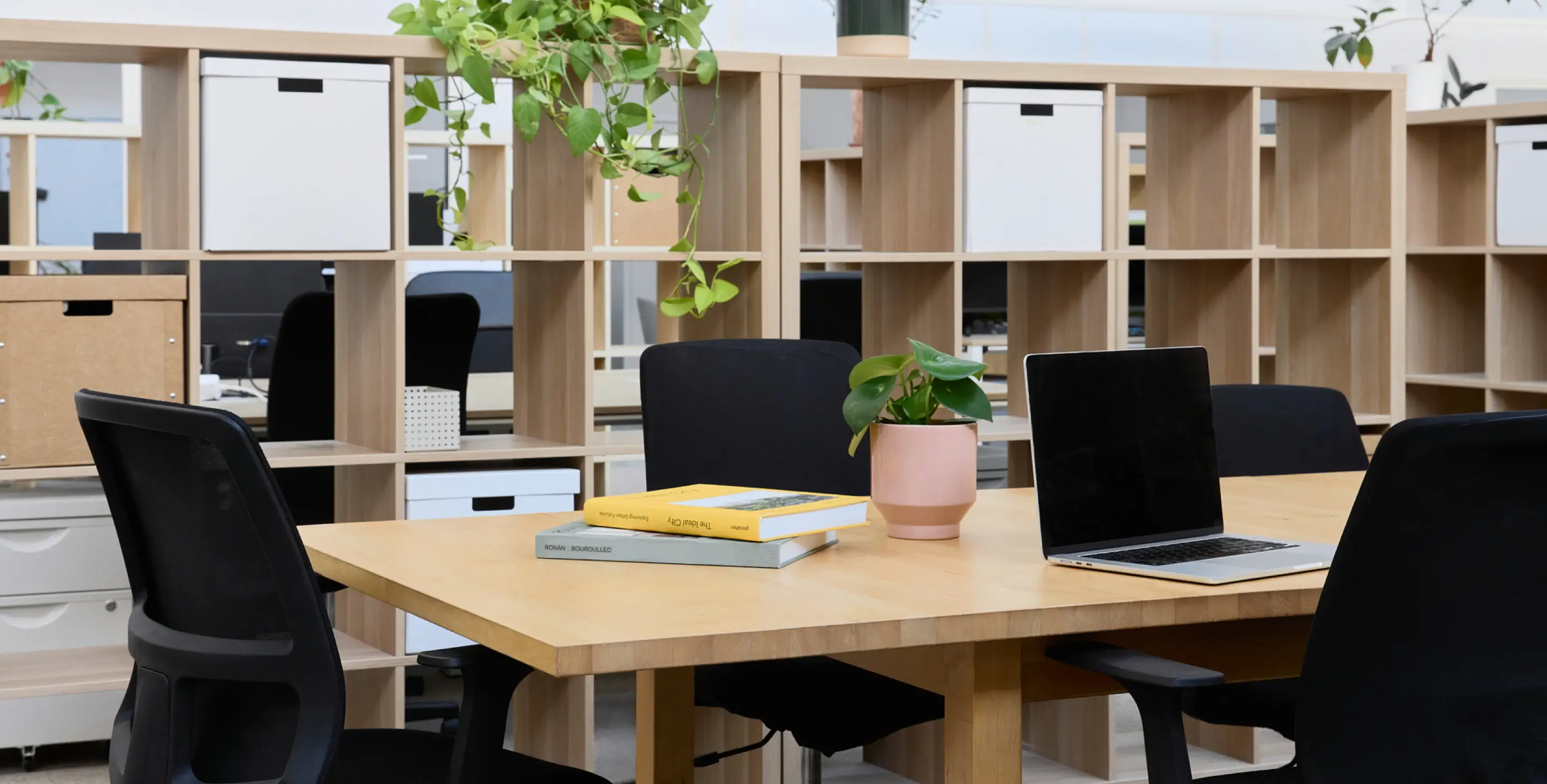 Bureau moderne au sein du coworking Greenspace avec une table en bois comportant un ordinateur portable, des livres et une plante en pot, entourée de chaises noires avec des étagères ouvertes en arrière-plan.