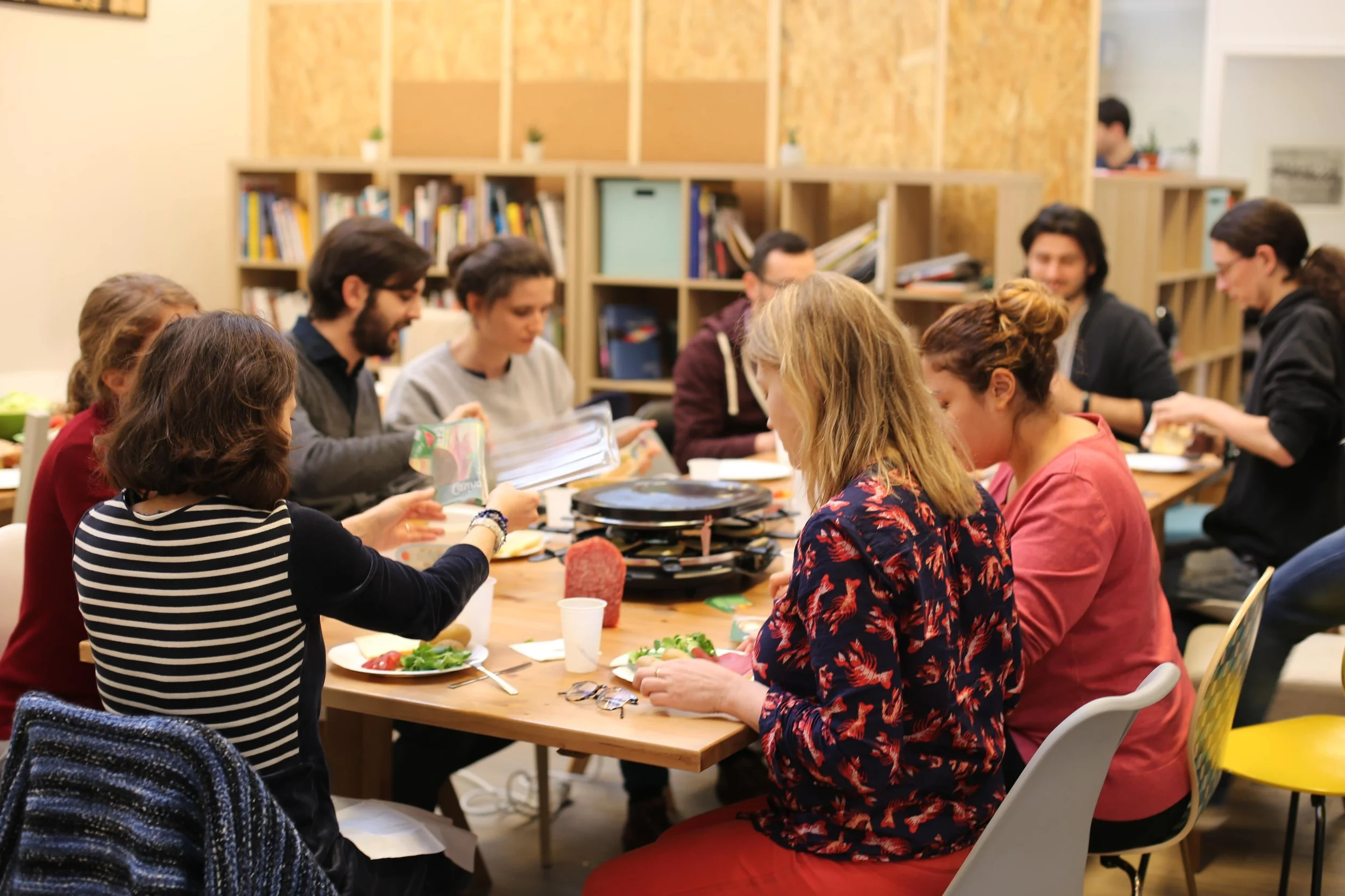 Groupe de personnes assises autour d'une table en bois partageant un repas dans une pièce avec des étagères remplies de livres.