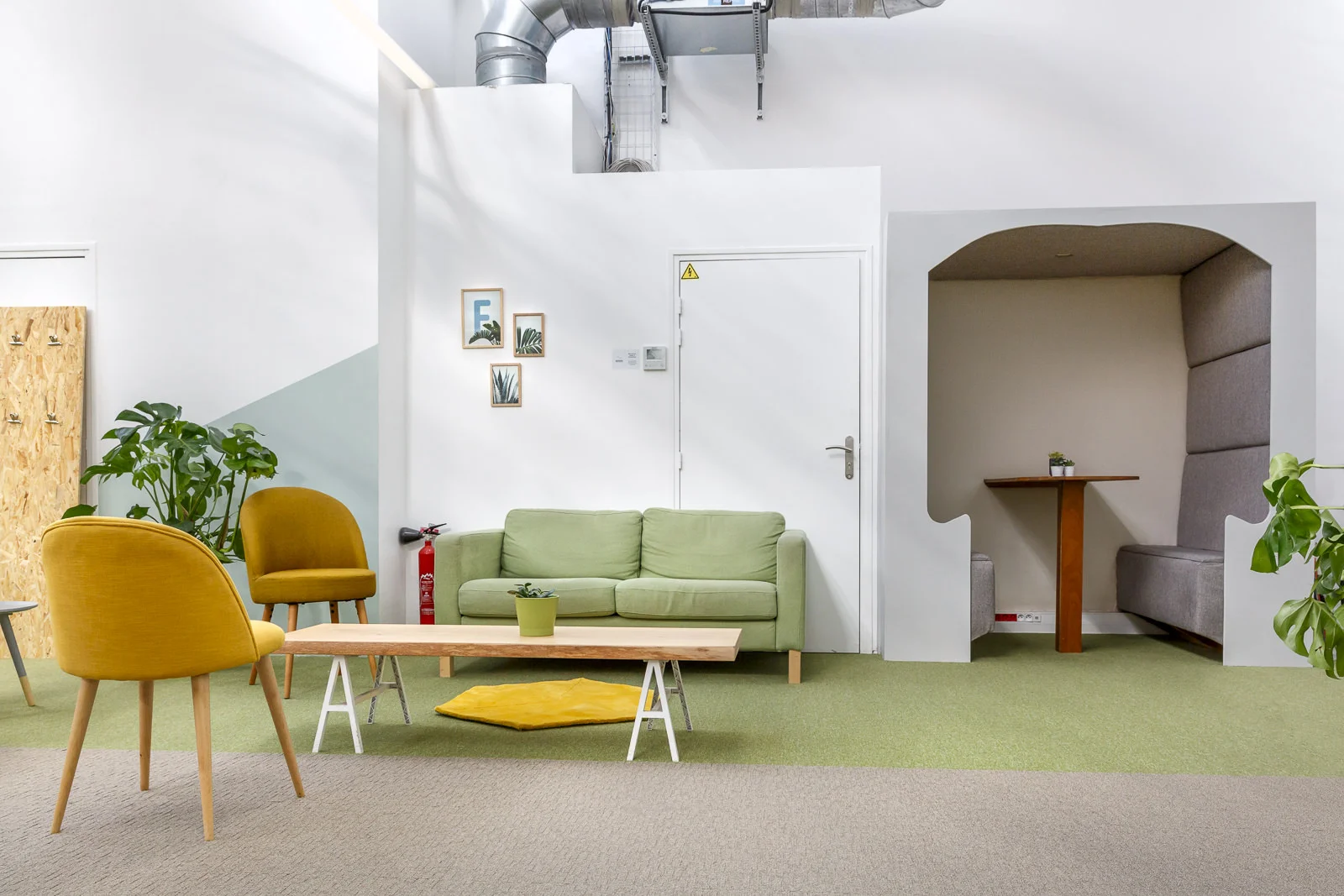Salle de repos lumineuse avec canapé vert, deux fauteuils jaunes, table basse en bois, plantes vertes, et une alcôve grise avec table et sièges intégrés.