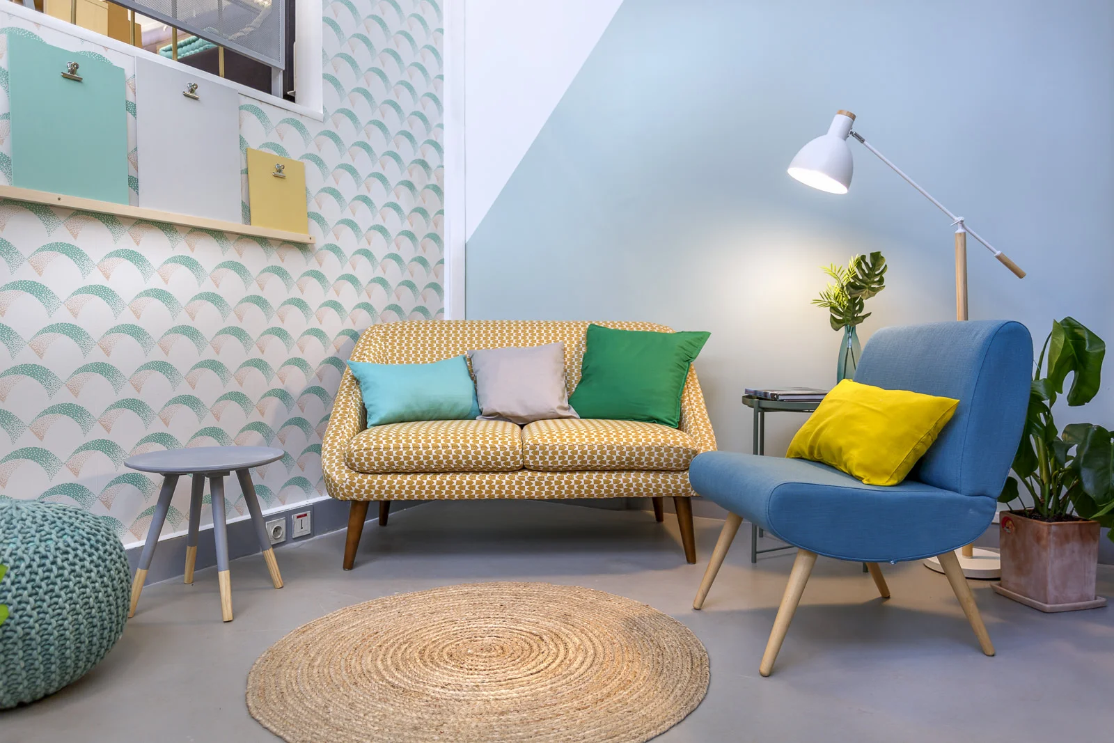 Salon moderne avec un fauteuil bleu, un canapé jaune à motifs, des coussins colorés, une lampe sur pied blanche et une plante verte.