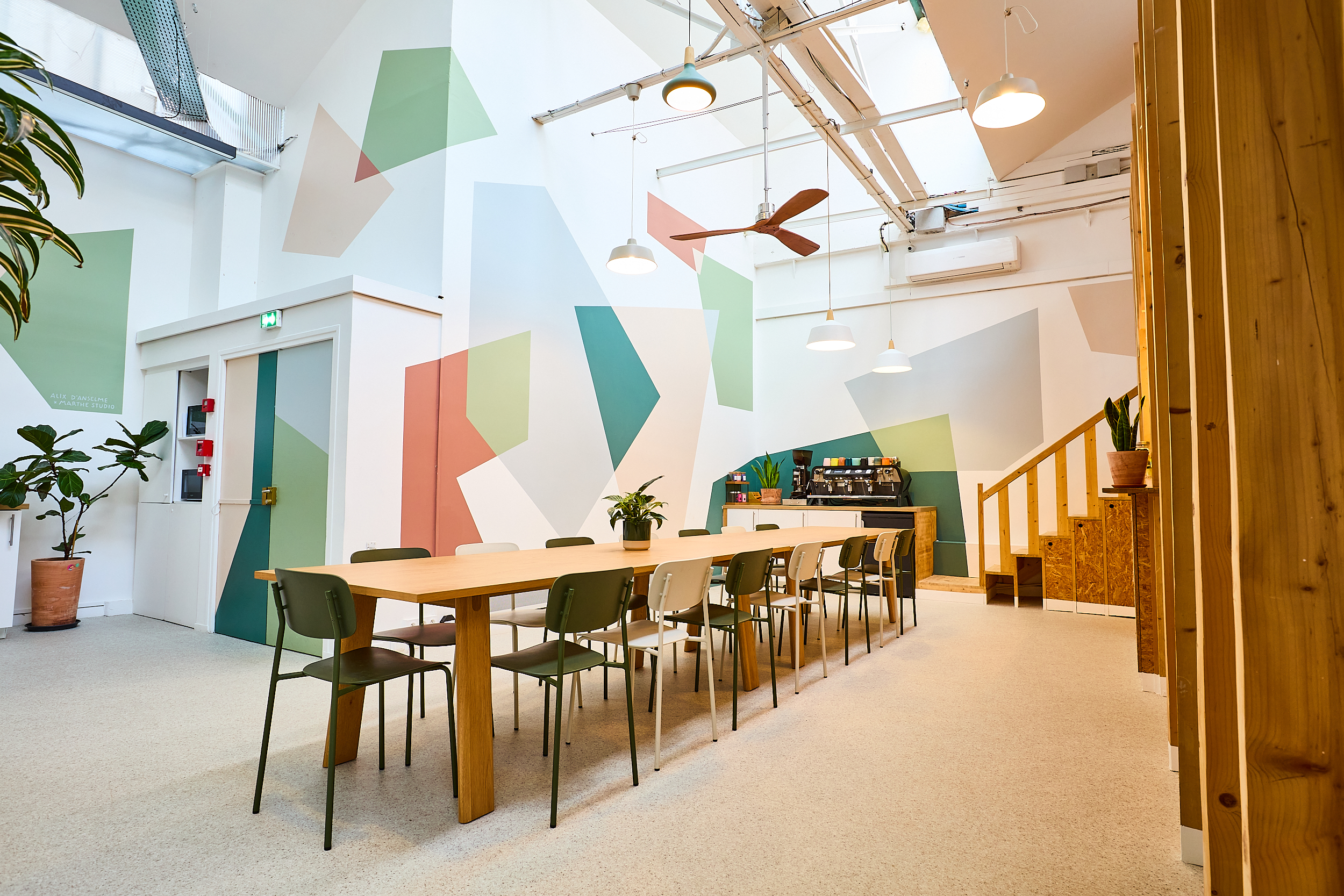 Cafétéria moderne avec grande table en bois, chaises vertes et blanches, murs colorés géométriques.