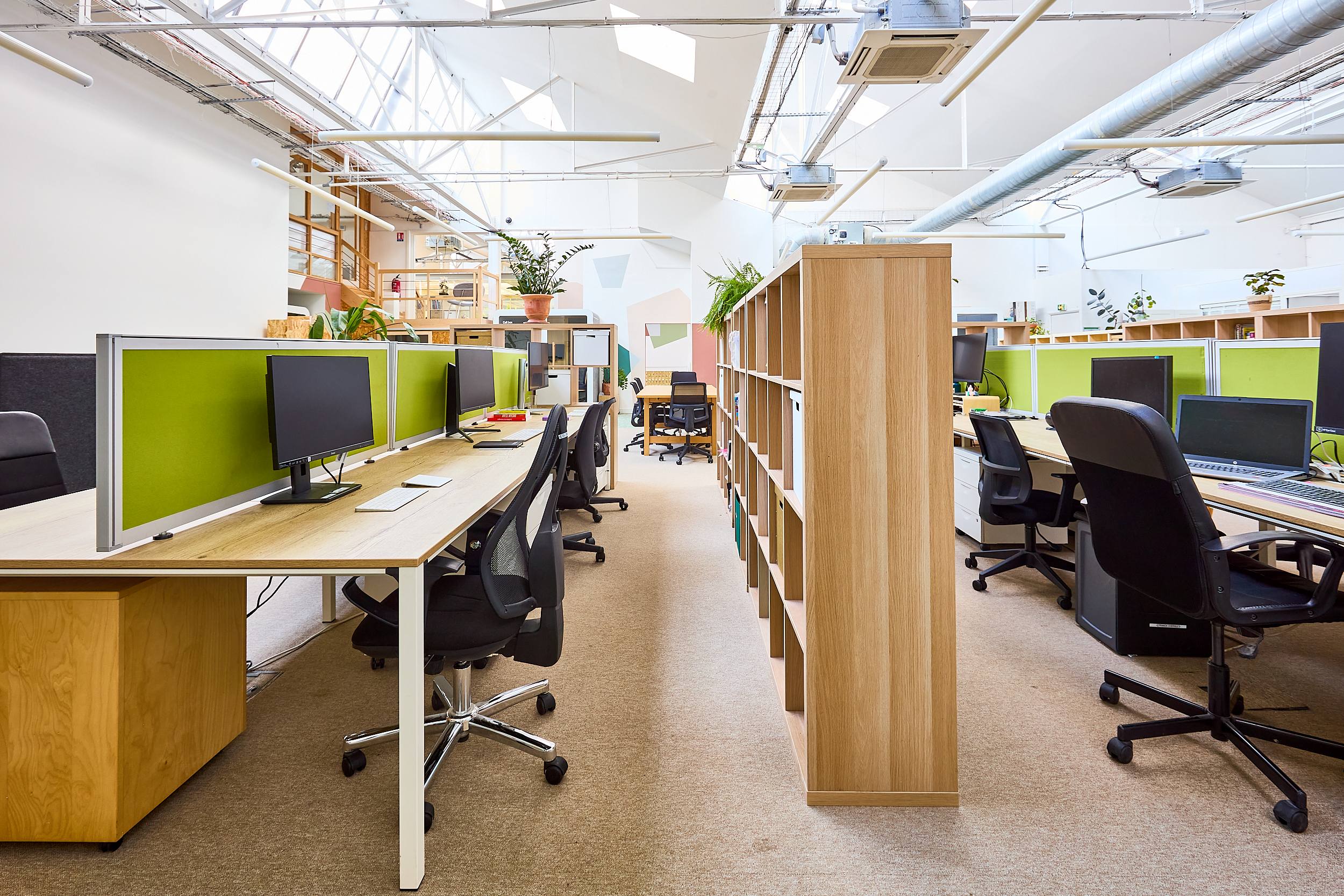 Espace de travail open space lumineux avec bureaux, chaises noires, écrans et séparateurs verts.