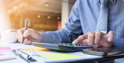 Person in a blue shirt and striped tie using a calculator and writing notes on paper at a desk with documents and a smartphone.