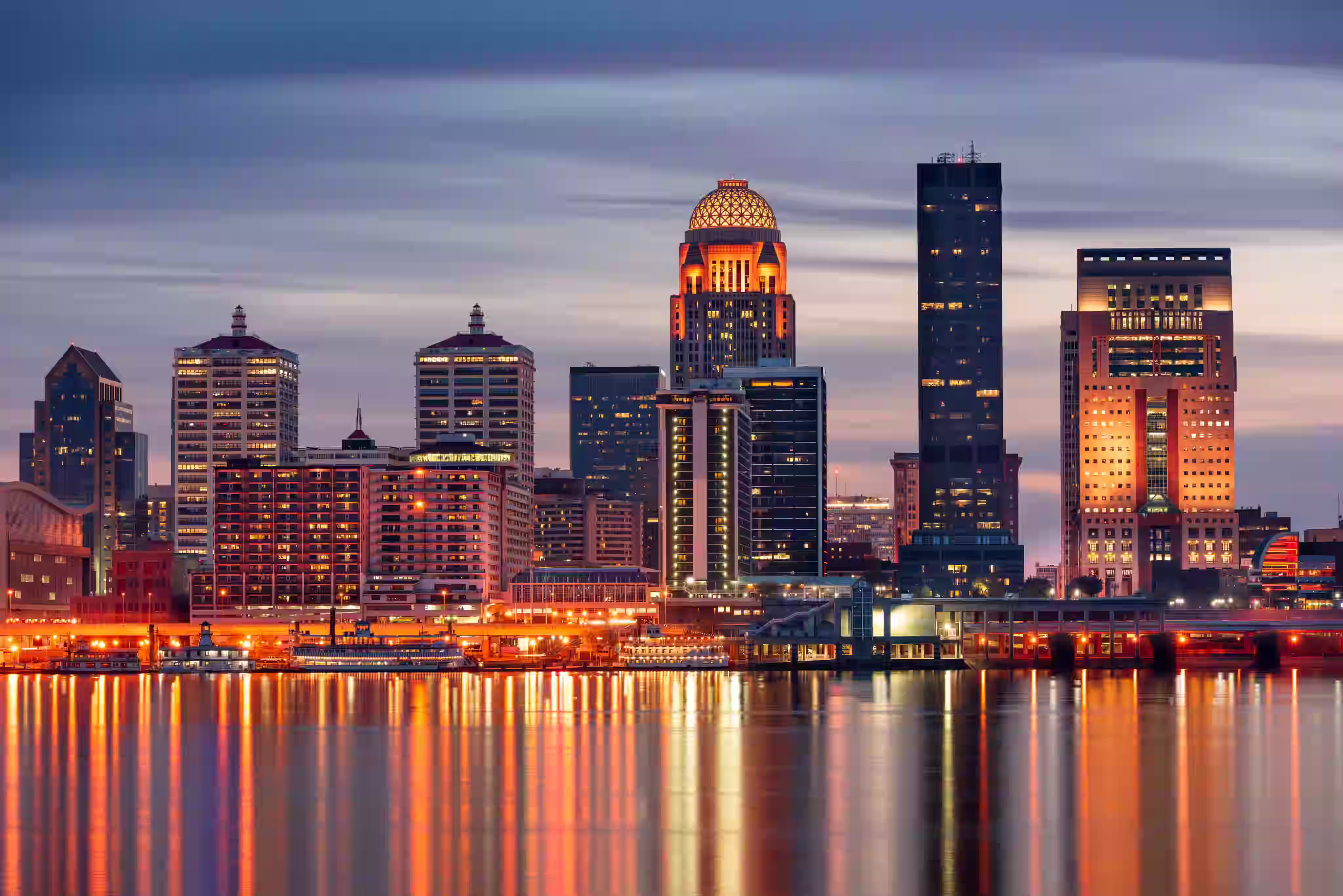 Evening skyline of Louisville, Kentucky, with illuminated buildings reflecting on the Ohio River.