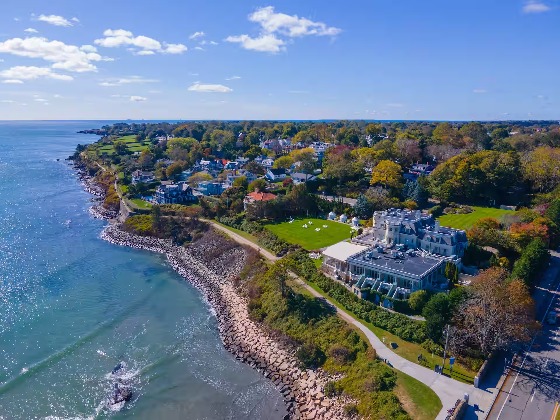 Aerial view of a coastal neighborhood with houses, green lawns, a walking path, and rocky shoreline under a blue sky with scattered clouds.