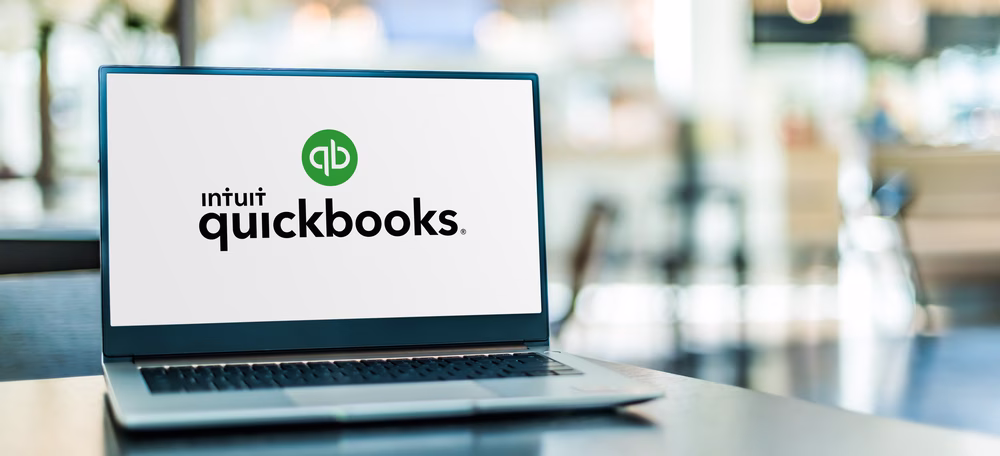 Laptop on a table displaying the Intuit QuickBooks logo on the screen.