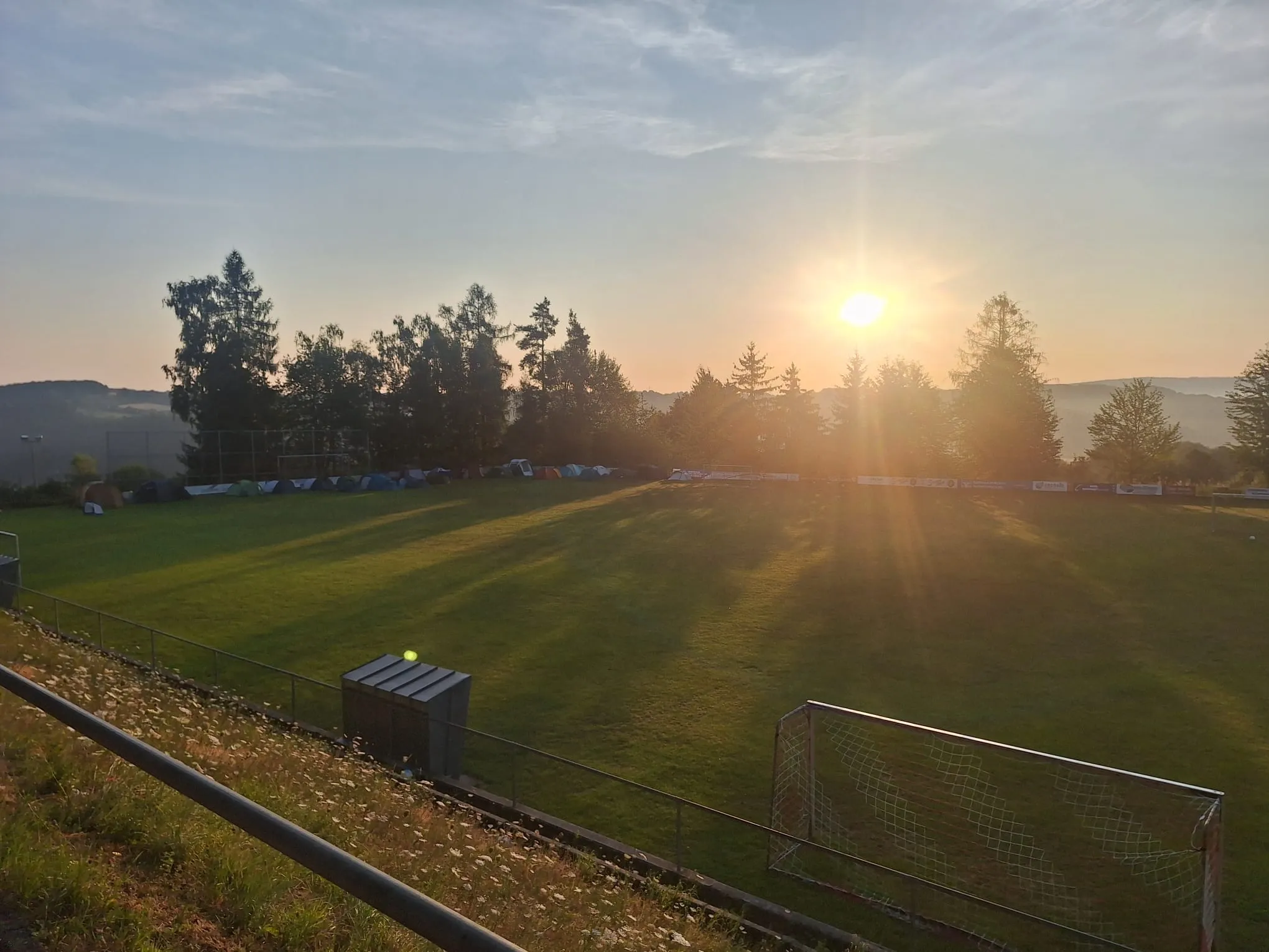 Sonnenuntergang über einem grasbewachsenen Fußballfeld mit einem Torpfosten und einer Reihe von Zelten in der Nähe von Bäumen.