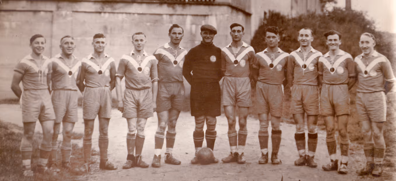 Schwarz-Weiß-Foto einer Fußballmannschaft in Trikots, die draußen in einer Reihe steht, mit einem Fußball davor, beschriftet mit „Fußball: Klub Mömbris: 1938”.