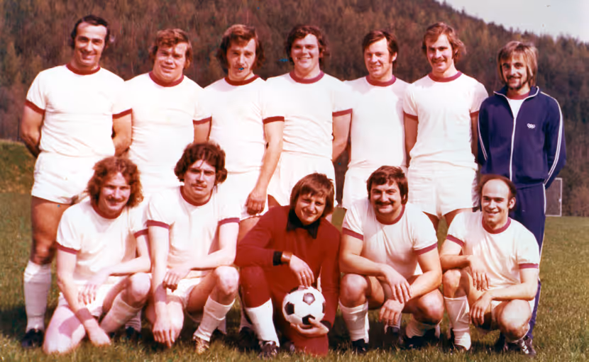 Die Fußballmannschaft der Männer von 1970 posiert im Freien auf einer Wiese vor einem bewaldeten Hügel im Hintergrund. Sie tragen weiße Trikots, mit Ausnahme eines Spielers in Rot, der einen Fußball in der Hand hält.
