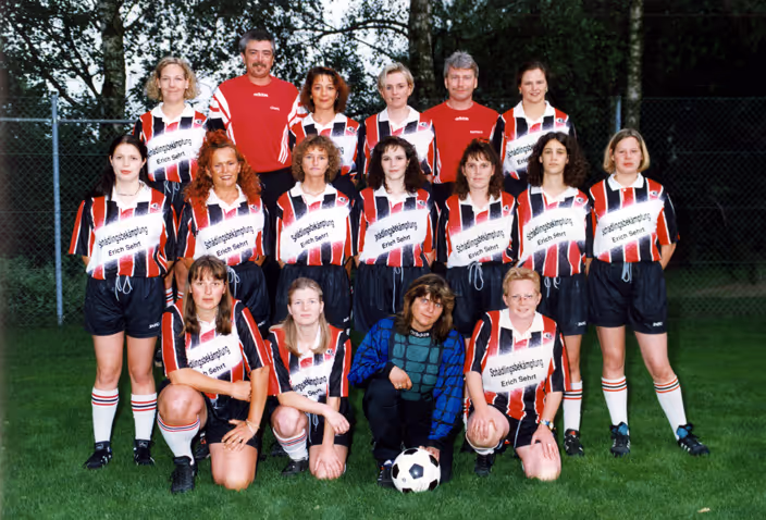Frauenfußballmannschaft in schwarz-weiß-rot gestreiften Trikots posiert auf grünem Rasen mit Trainer in rotem Hemd im Hintergrund und Torhüterin in blau-schwarzem Outfit, die neben einem Fußball kniet.