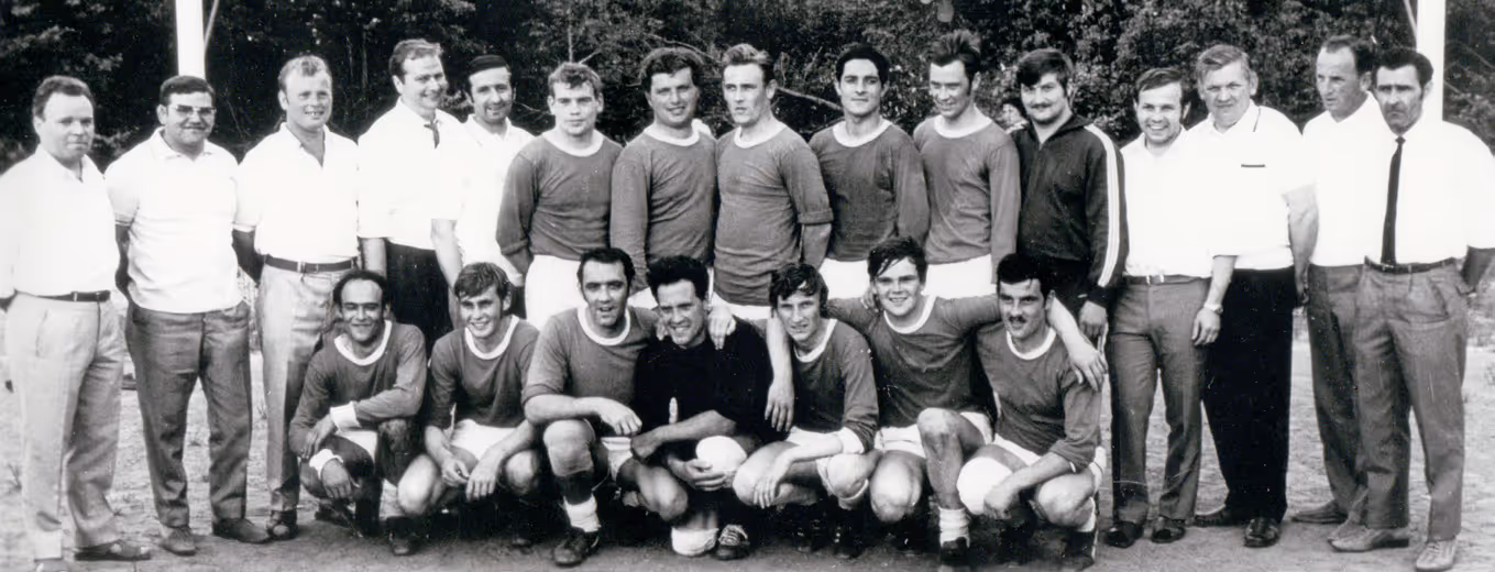 Schwarz-Weiß-Foto einer alten Fußballmannschaft mit vierzehn Spielern in Uniform und acht Männern in Freizeit- oder formeller Kleidung, die hinter ihnen auf einem Feld stehen.