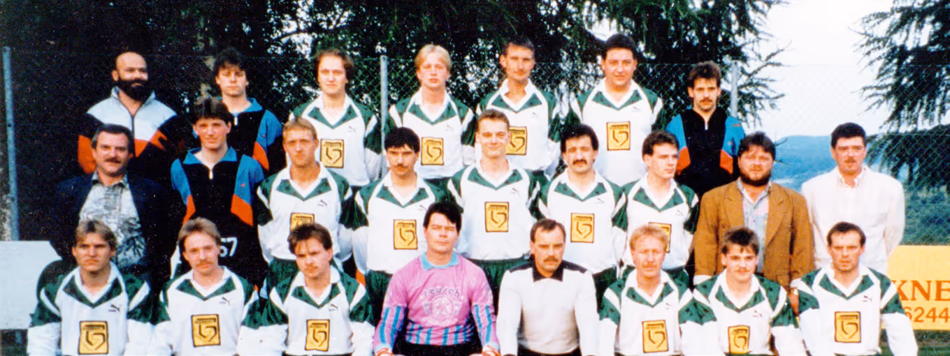 Gruppenfoto einer Männerfußballmannschaft in weiß-grünen Trikots, wobei zwei Männer vorne andere Shirts tragen und im Freien stehen und sitzen.