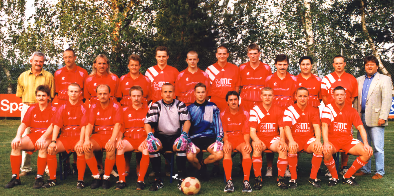 Eine Fußballmannschaft bestehend aus 20 Männern, die im Freien vor Bäumen posieren, wobei die Spieler rote Trikots tragen und zwei Torhüter in auffälligen Trikots mit einem Fußball im Vordergrund sitzen.