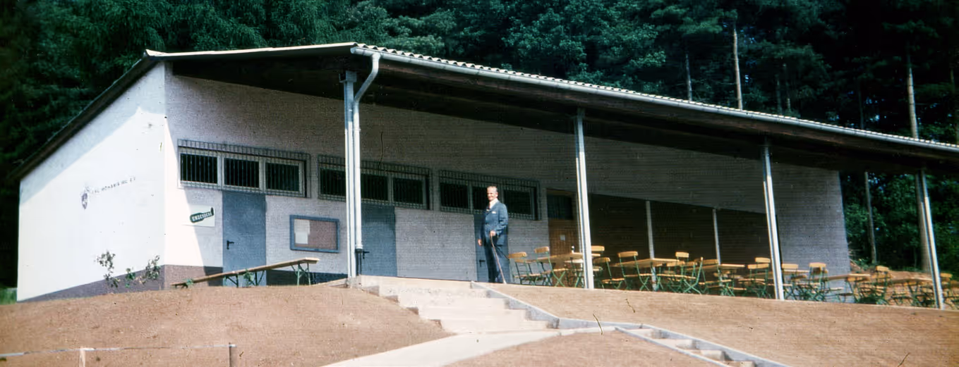 Einstöckiges weißes Gebäude mit einem Mann davor, umgeben von Tischen und Stühlen im Freien und einem Wald im Hintergrund.