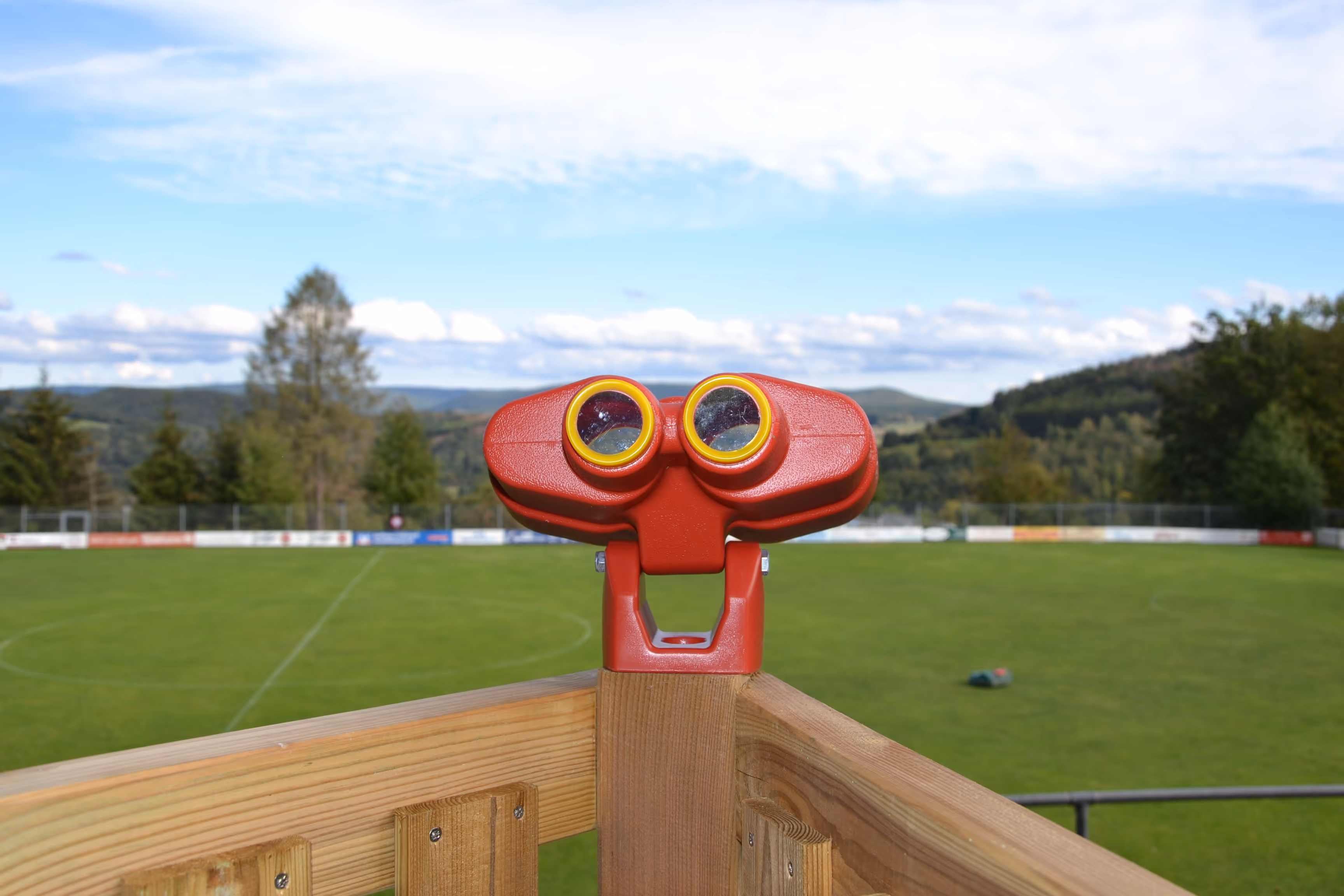 Rotes Fernglas, montiert an einem Holzgeländer mit Blick auf einen grünen Sportplatz, im Hintergrund Bäume und Hügel.