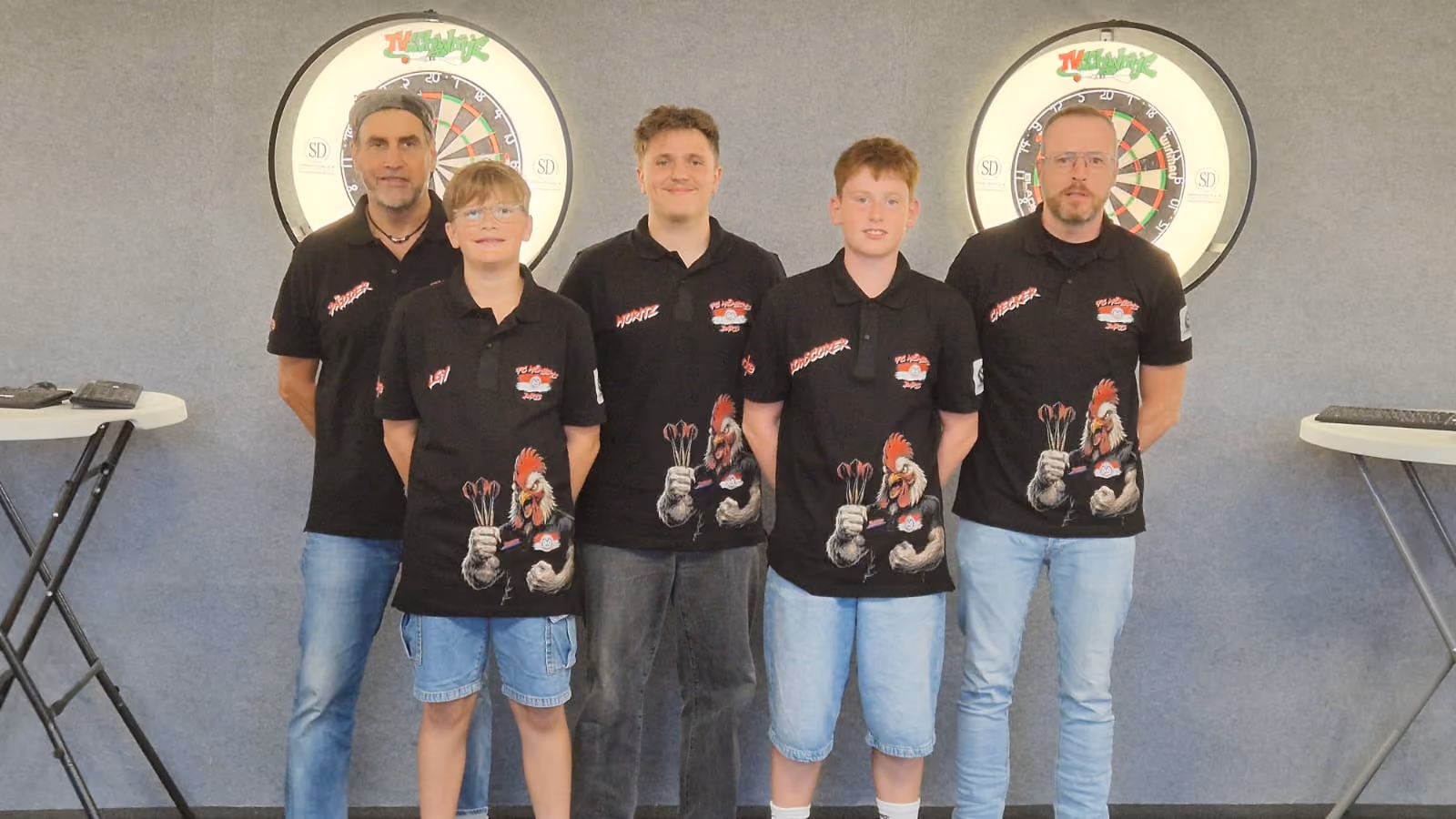 Fünf Personen mit schwarzen Poloshirts mit einem Hahn-Logo und Darts-Pfeilen stehen vor zwei Dartscheiben an einer Wand.