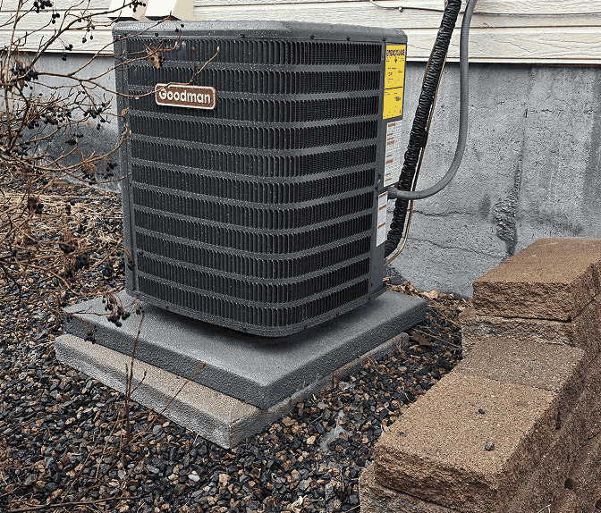 Goodman air conditioning unit installed on a concrete slab outside a house with gravel and brick steps nearby.