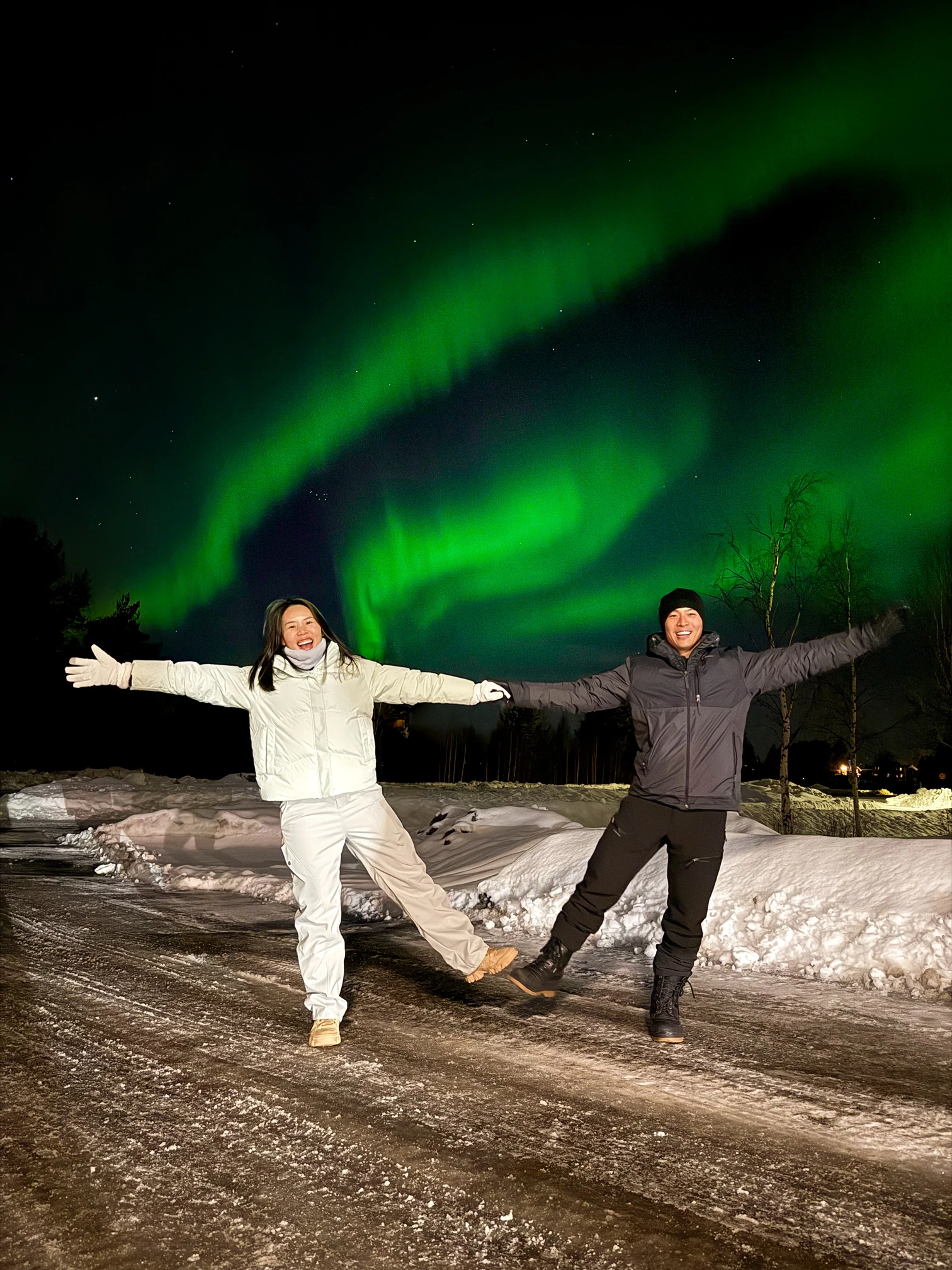 Two people holding hands and smiling with arms outstretched under green northern lights at night in snowy landscape.