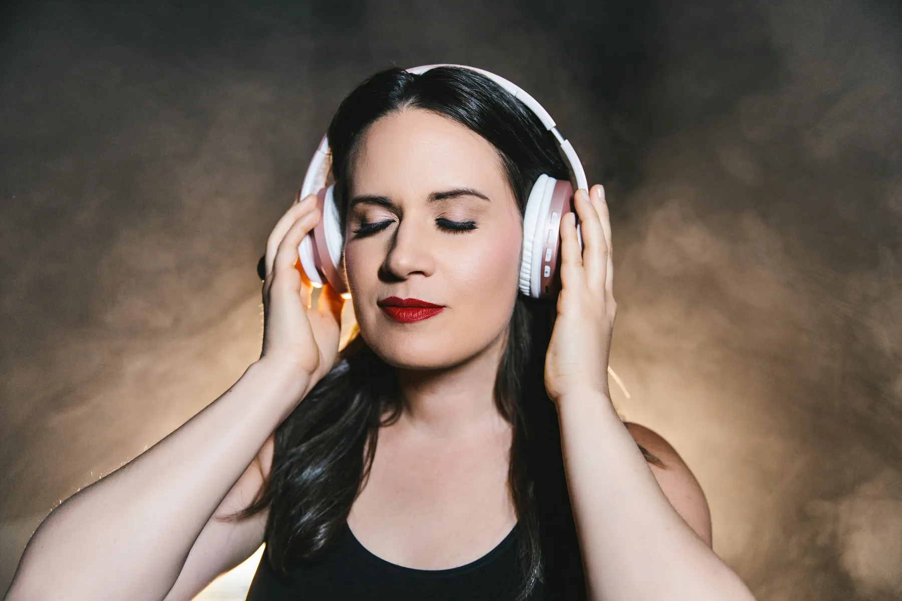 Woman with closed eyes wearing white headphones and enjoying music, with a dark smoky background.