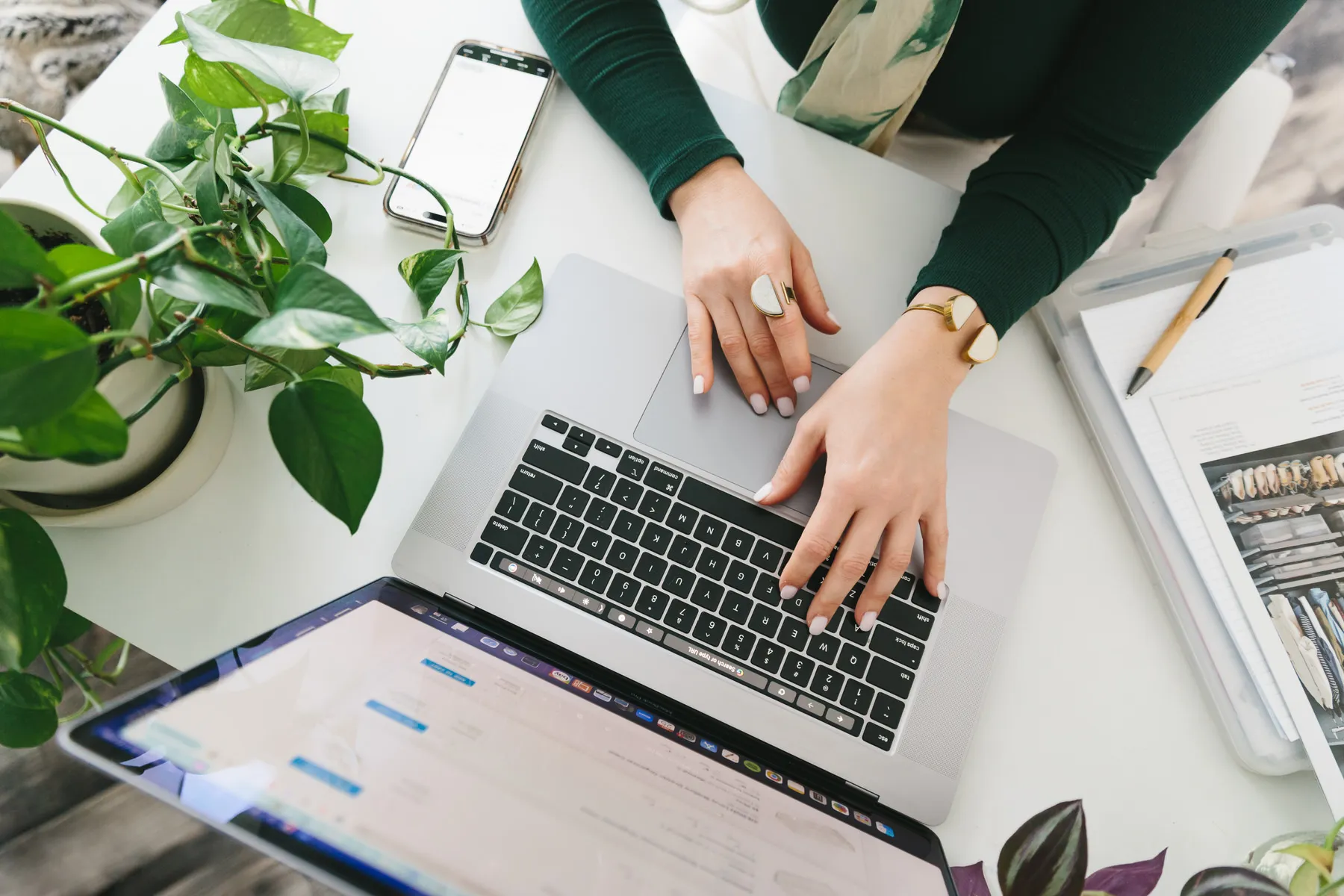 Person typing on a laptop keyboard at a white desk with plants, a smartphone, and a notebook nearby.