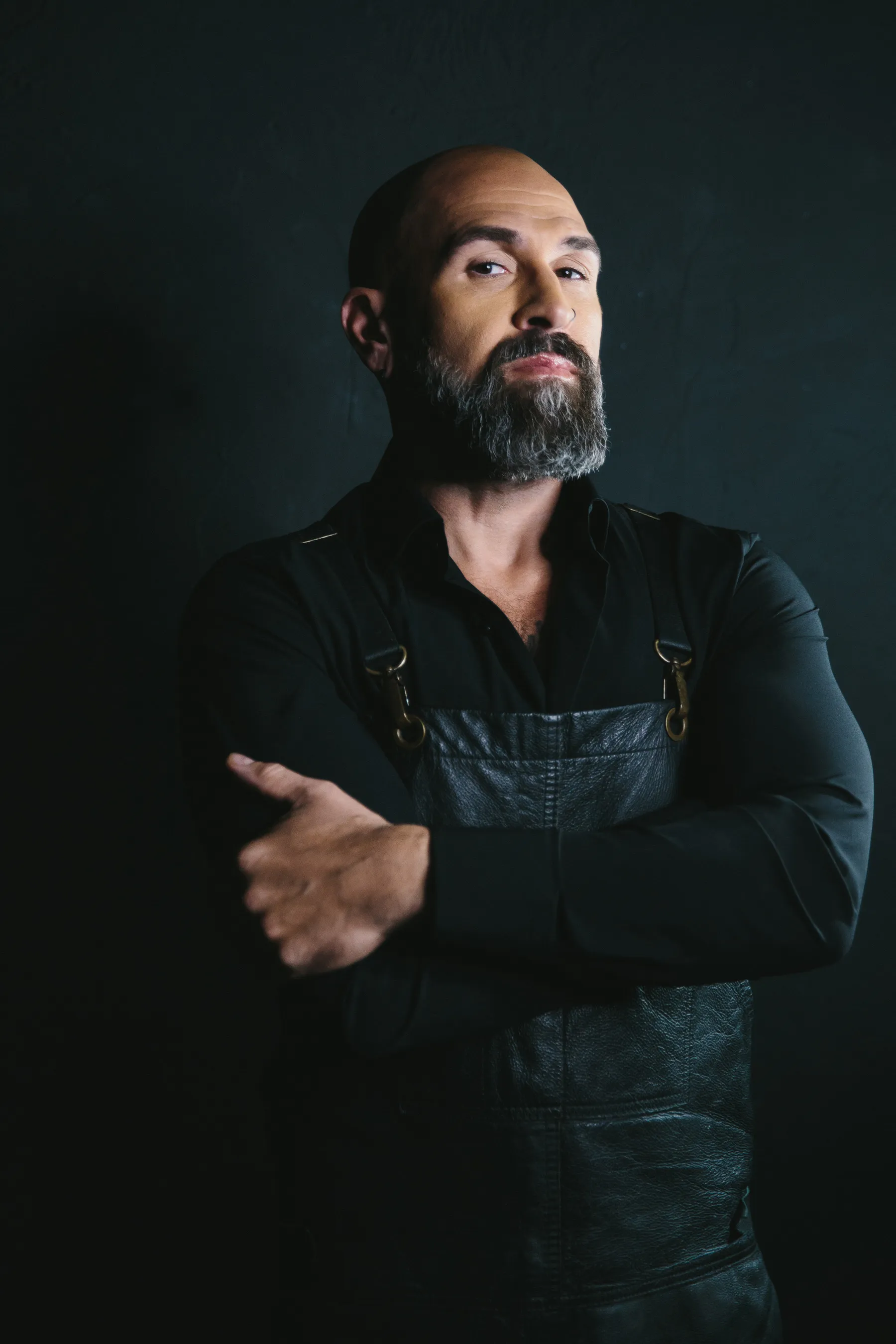 Confident bald man with a gray beard wearing a black shirt and leather apron against a dark background.