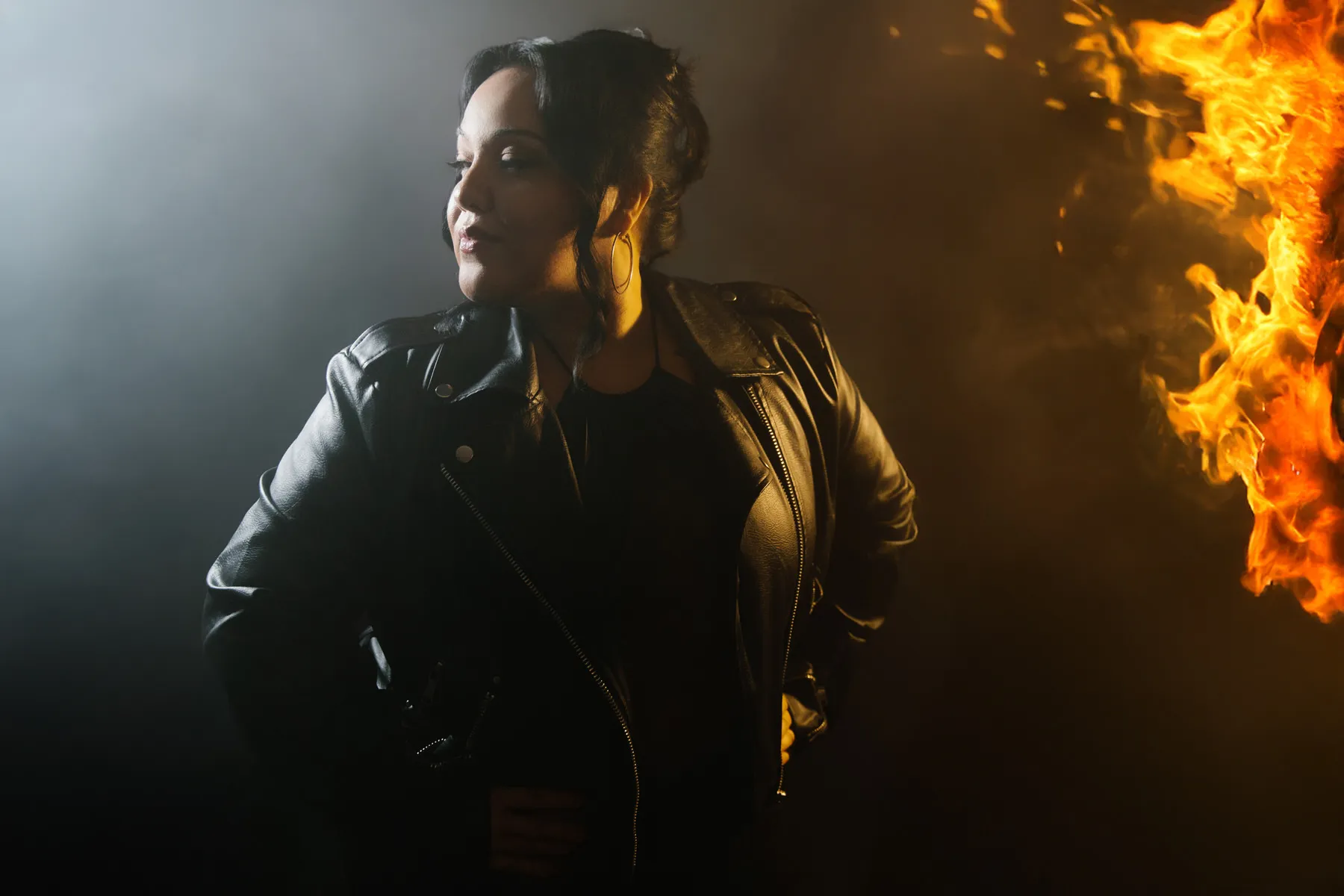 Woman in a black leather jacket standing with hands on hips looking sideways, with flames burning on the right side.