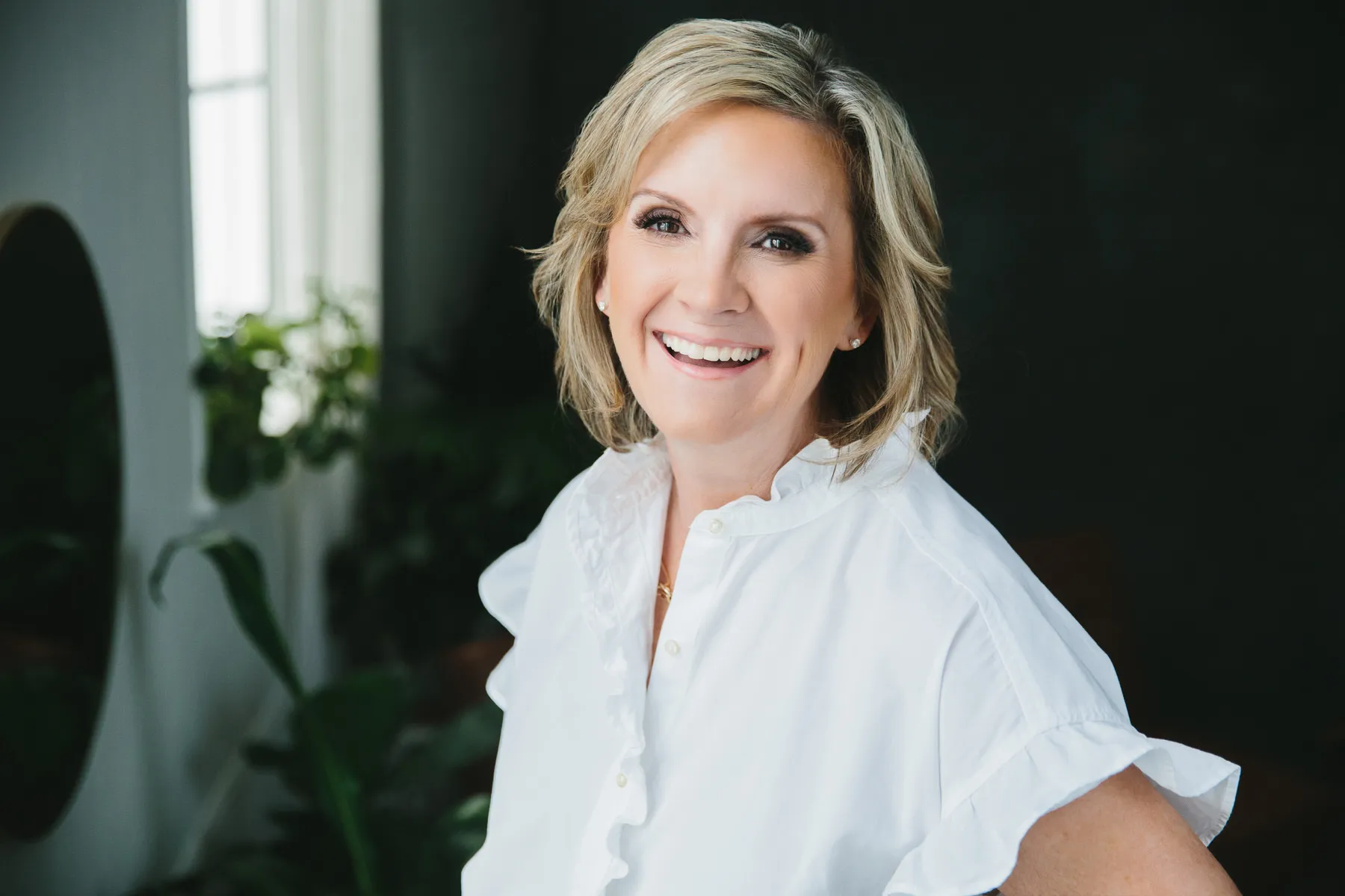 Smiling blonde woman wearing a white blouse standing indoors with plants and a dark background.