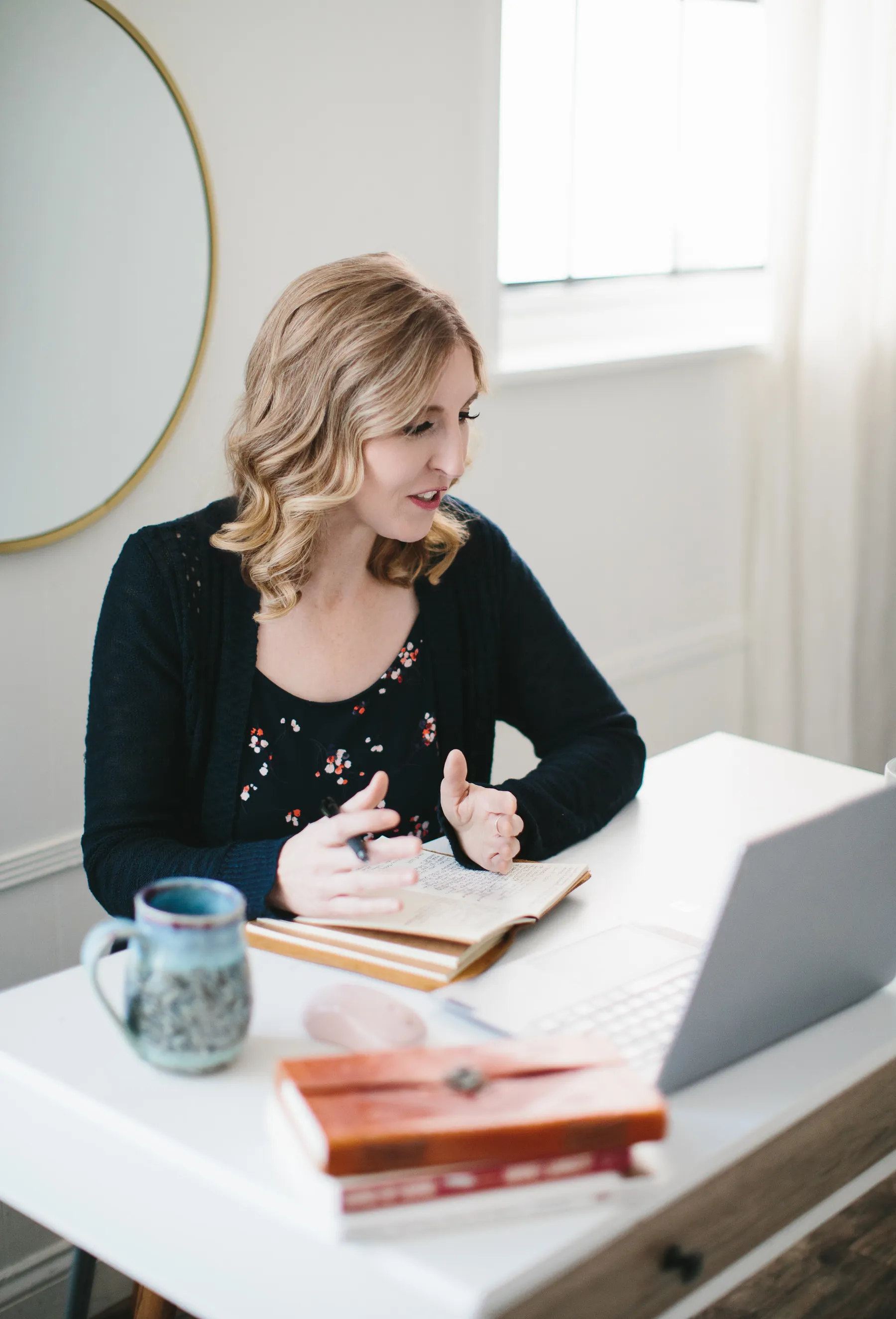 Woman with curled blonde hair discussing while sitting at a desk with a notebook, laptop, and ceramic mug.