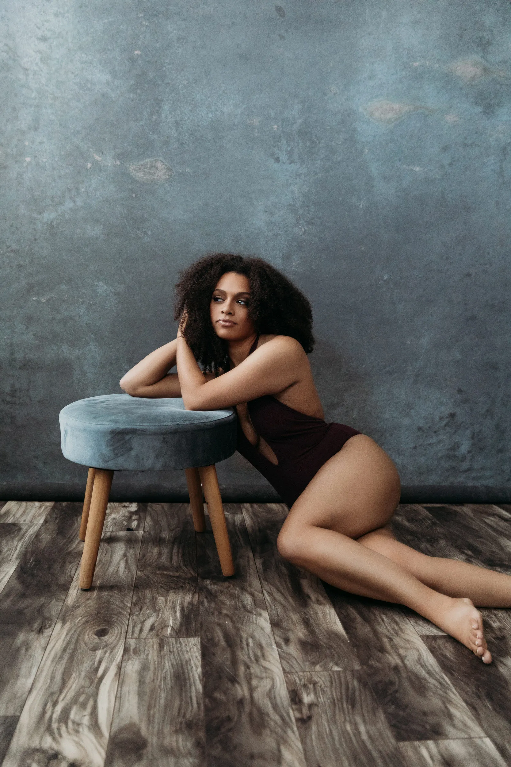 Woman with curly hair in a dark swimsuit leaning on a blue cushioned stool on a wood floor with a gray textured backdrop.