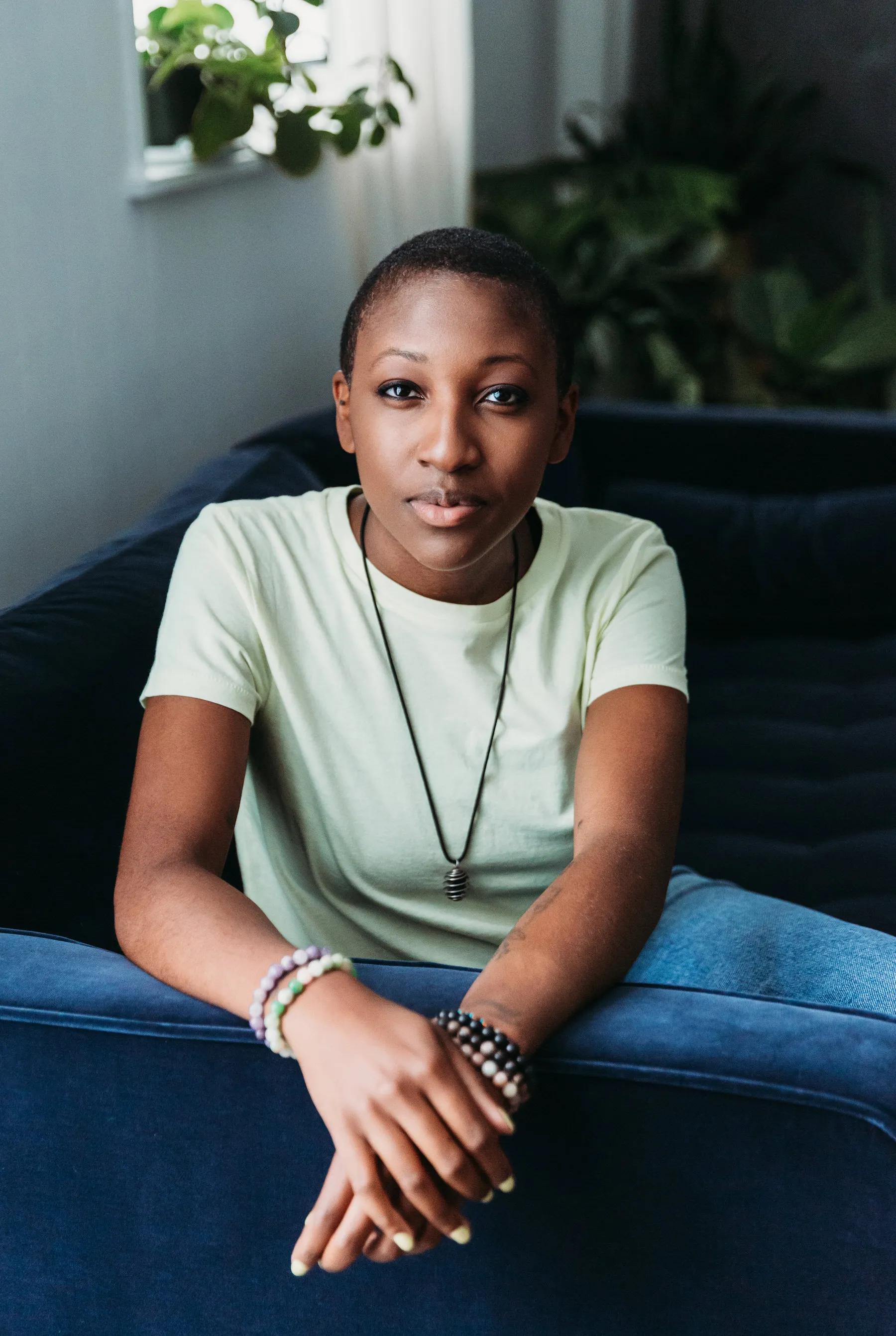 Young person wearing a light yellow t-shirt and bracelets, sitting on a blue couch with hands crossed and looking directly at the camera.