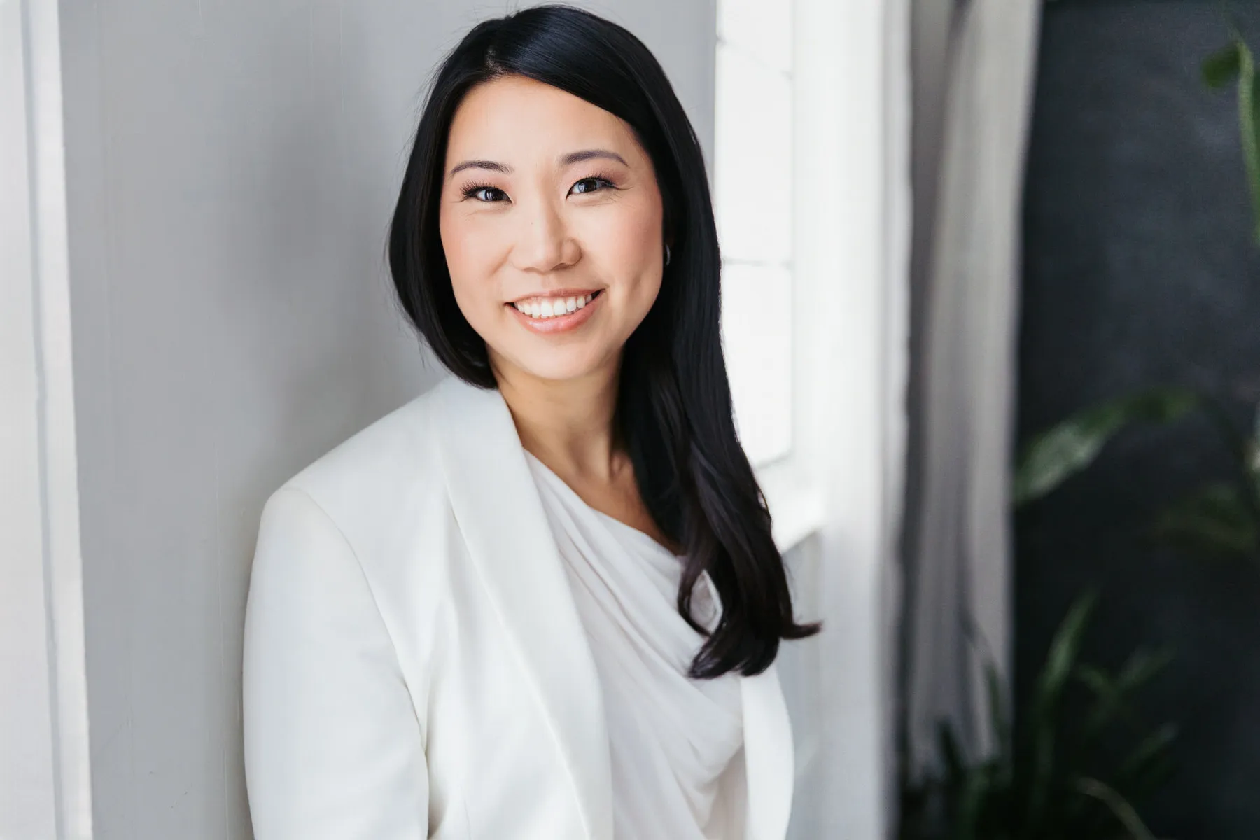 Smiling woman with long black hair wearing a white blazer and top, standing indoors near a window.