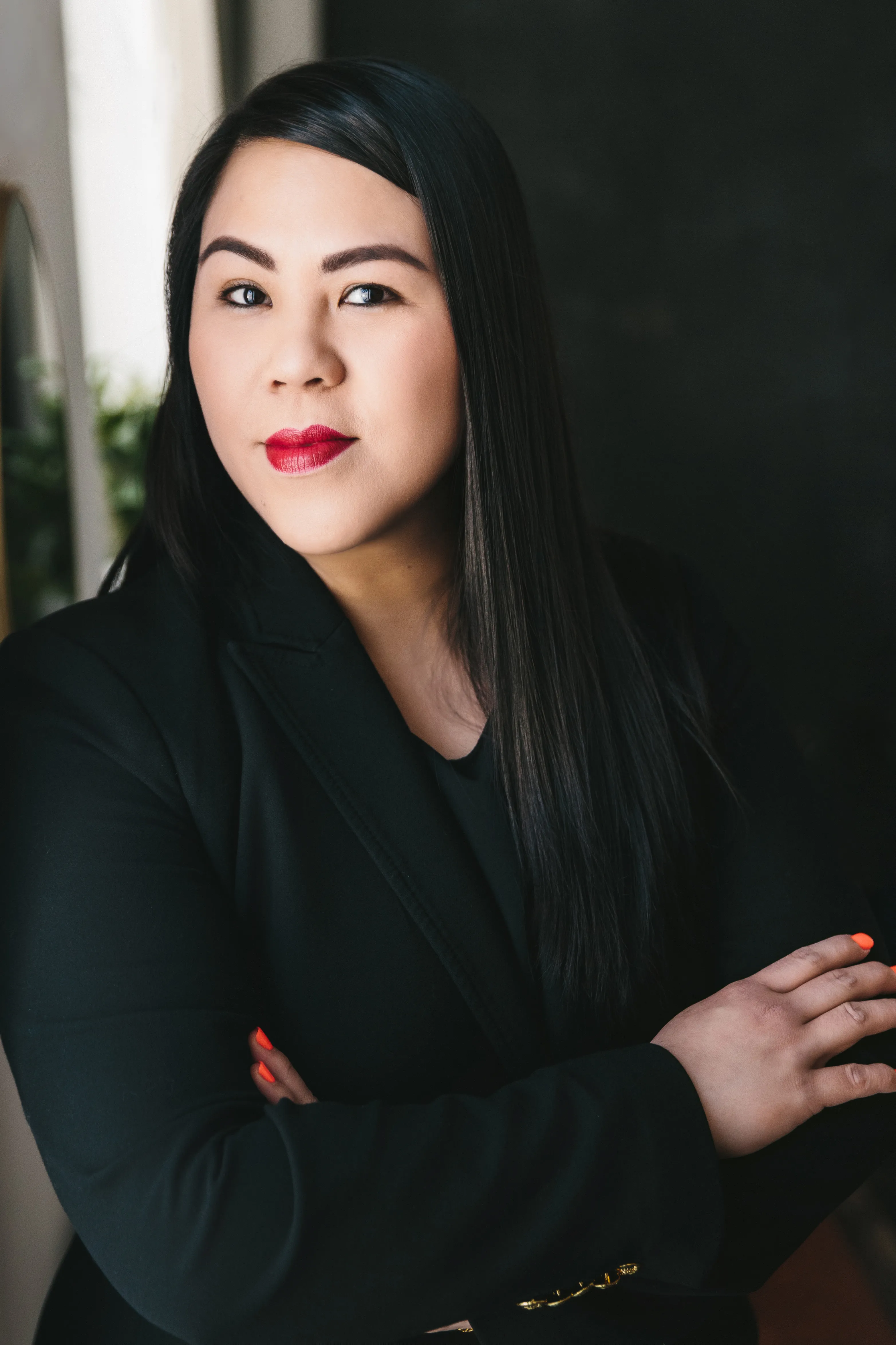 Confident woman with long black hair wearing a black blazer and red lipstick, arms crossed.