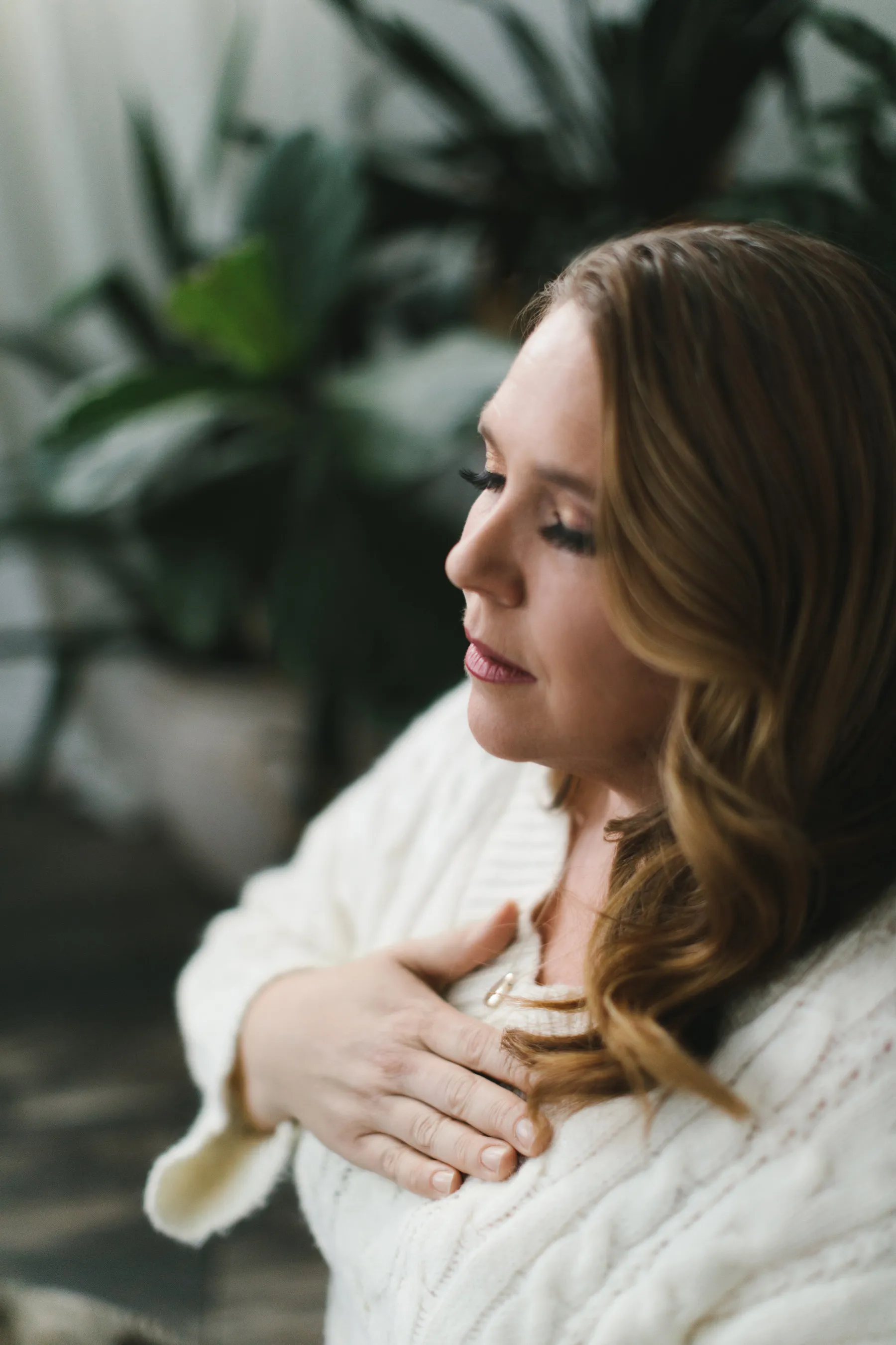 Woman with closed eyes, wearing a white sweater, resting her hand on her chest in a calm, indoor setting with blurred plants in the background.