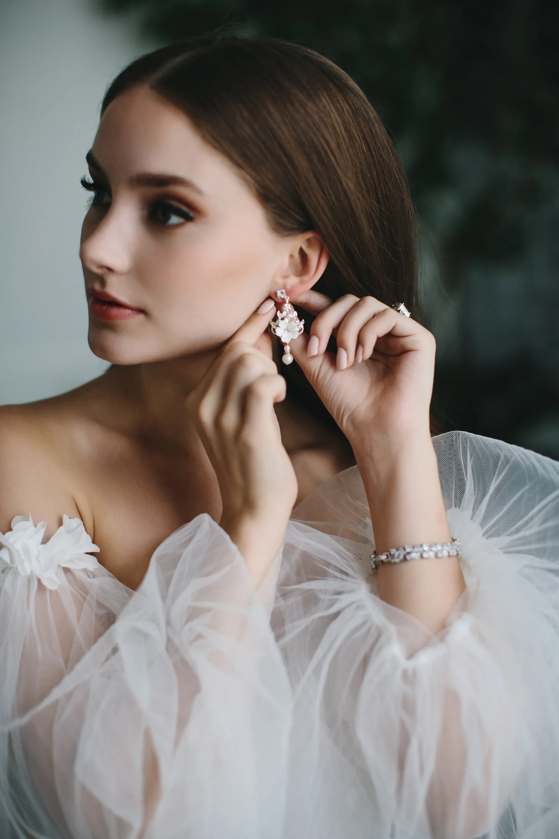 Young woman wearing a sheer white off-shoulder dress adjusts a floral pearl earring.