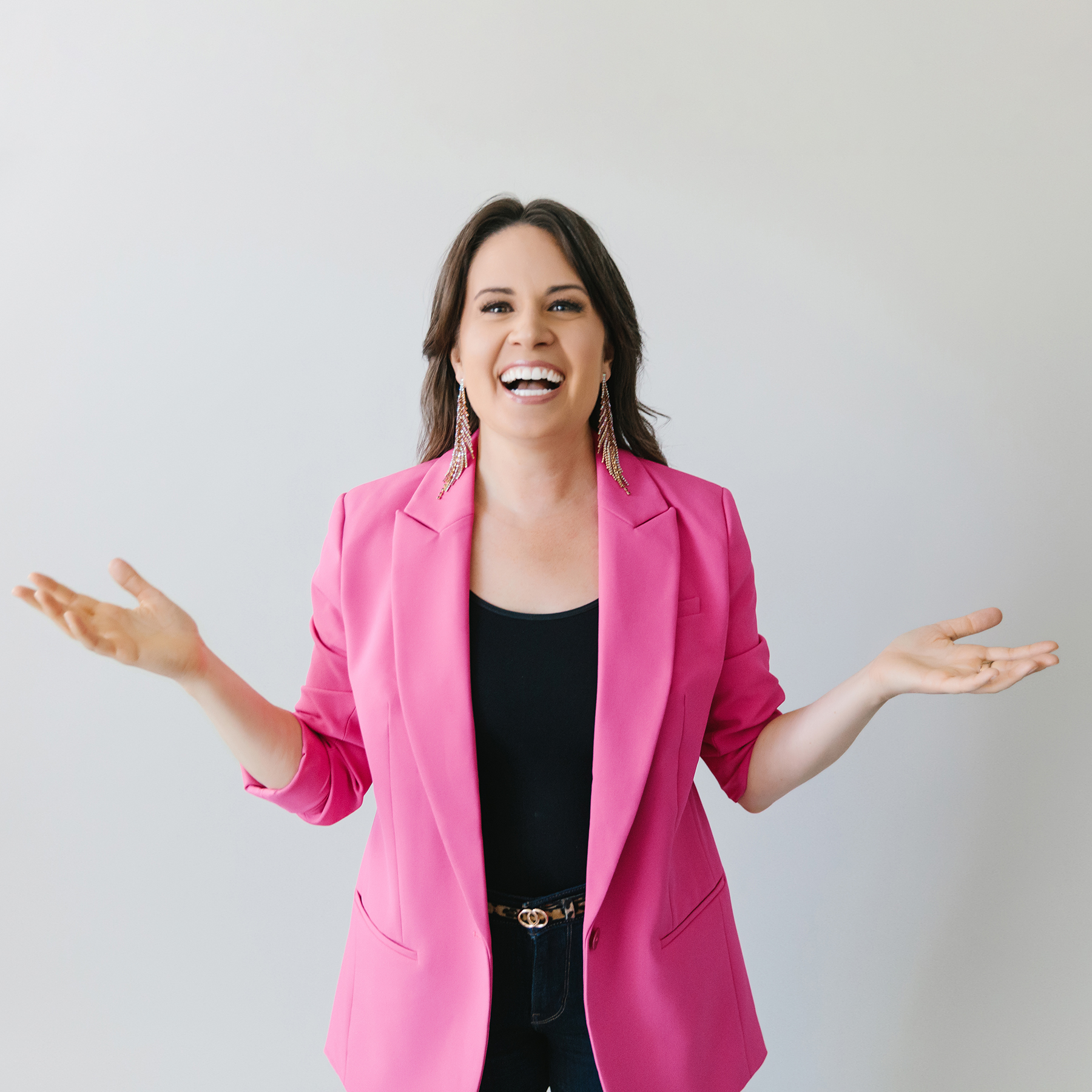 Smiling woman with dark hair wearing a bright pink blazer and large earrings, gesturing with open hands against a plain light background.