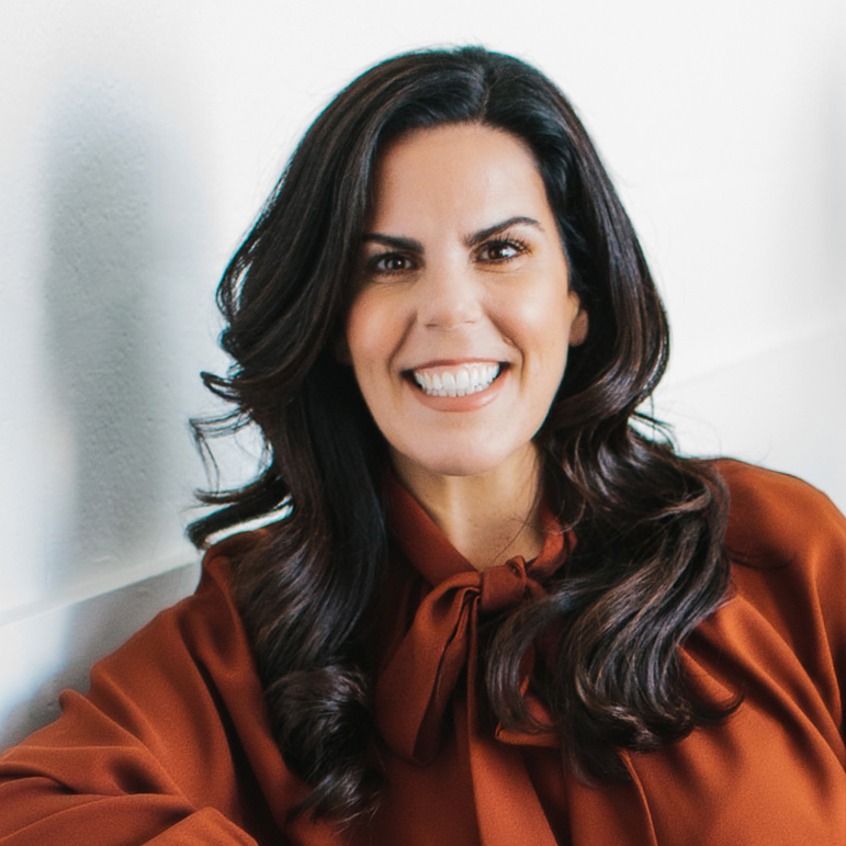 Smiling woman with long dark hair wearing a rust-colored blouse with a bow tie collar.