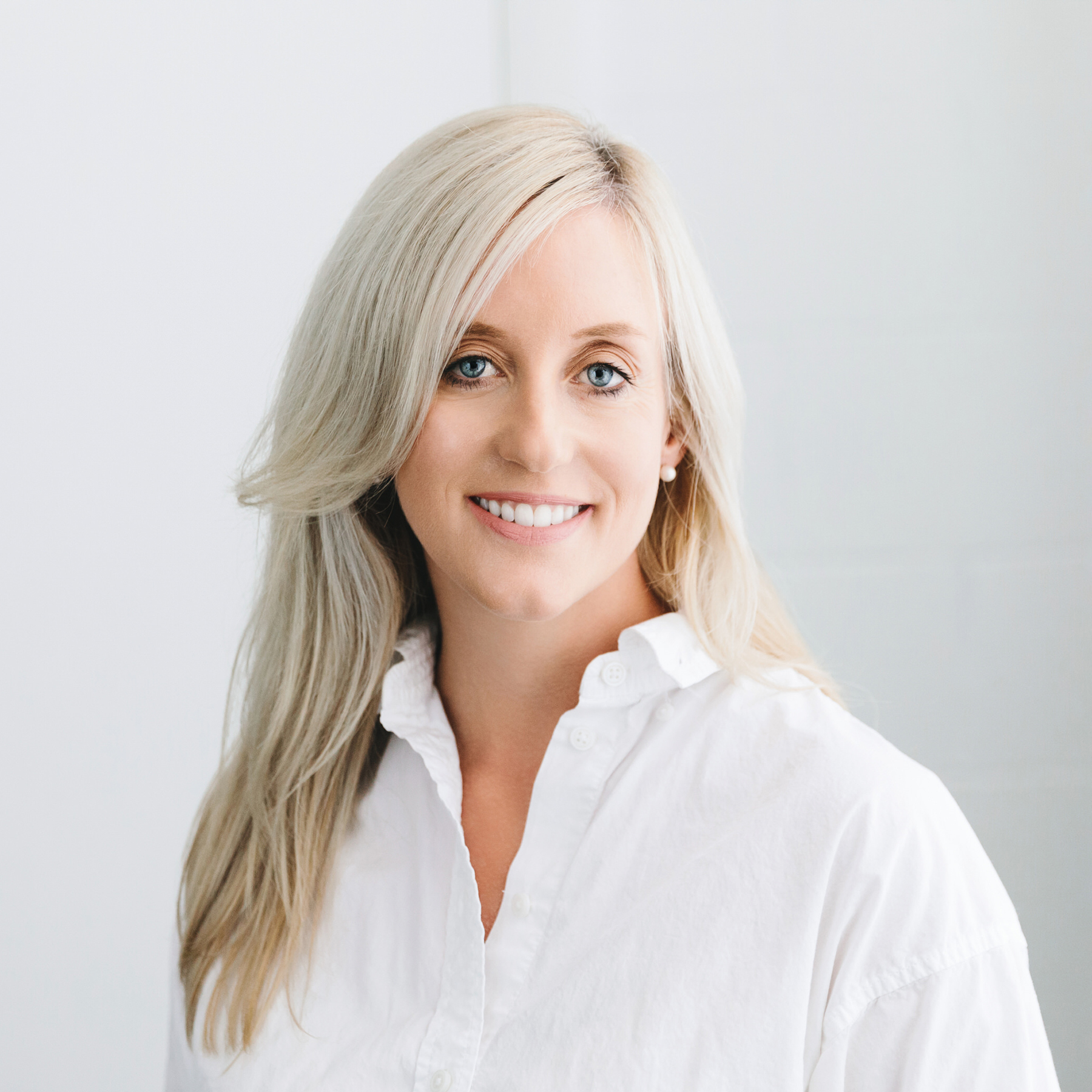 Smiling woman with long blonde hair wearing a white button-up shirt against a light background.